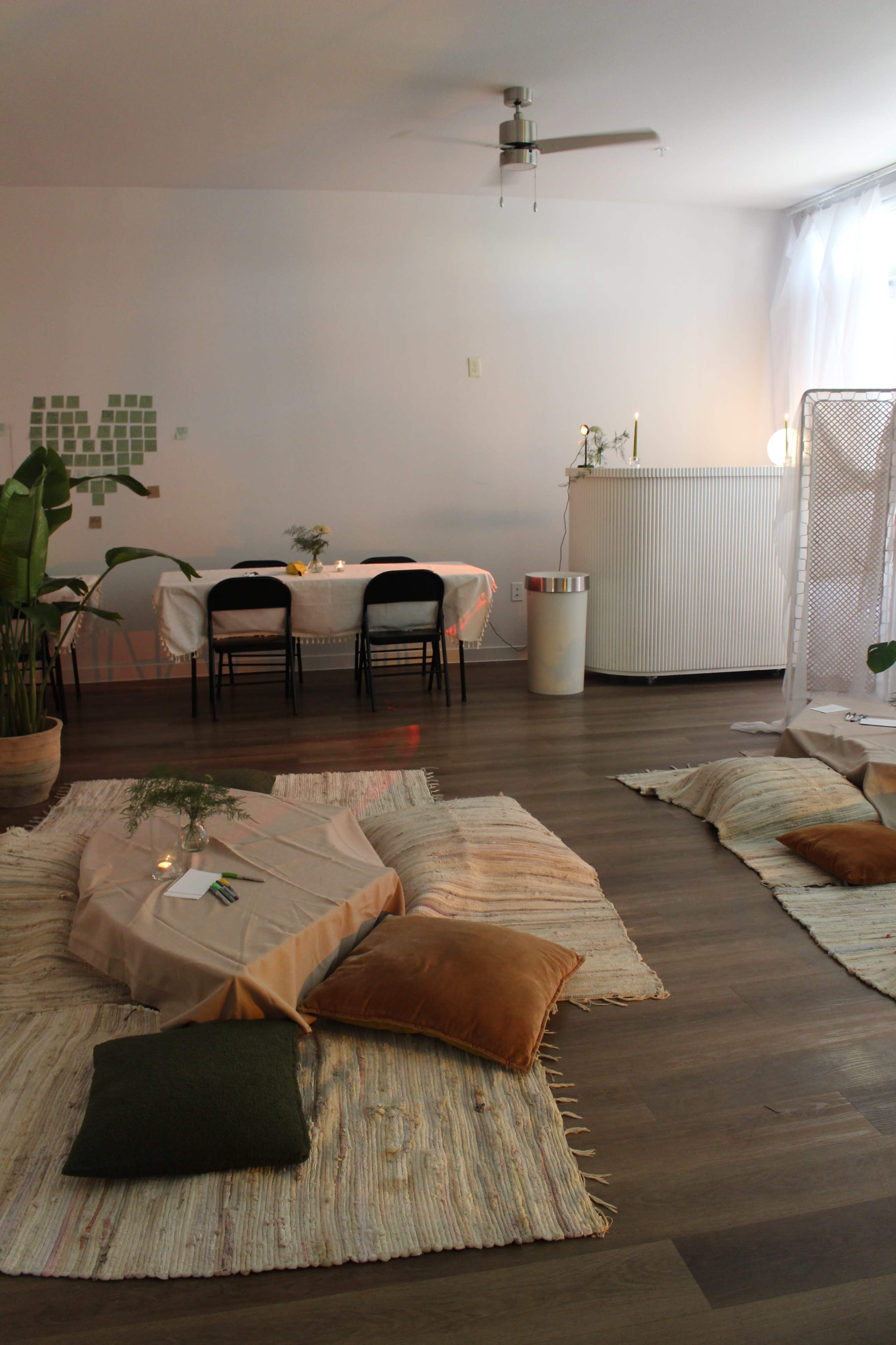 The image shows a serene indoor setting with a table and two chairs, surrounded by cushions and soft textiles on the floor, along with plants and a fan on the ceiling.