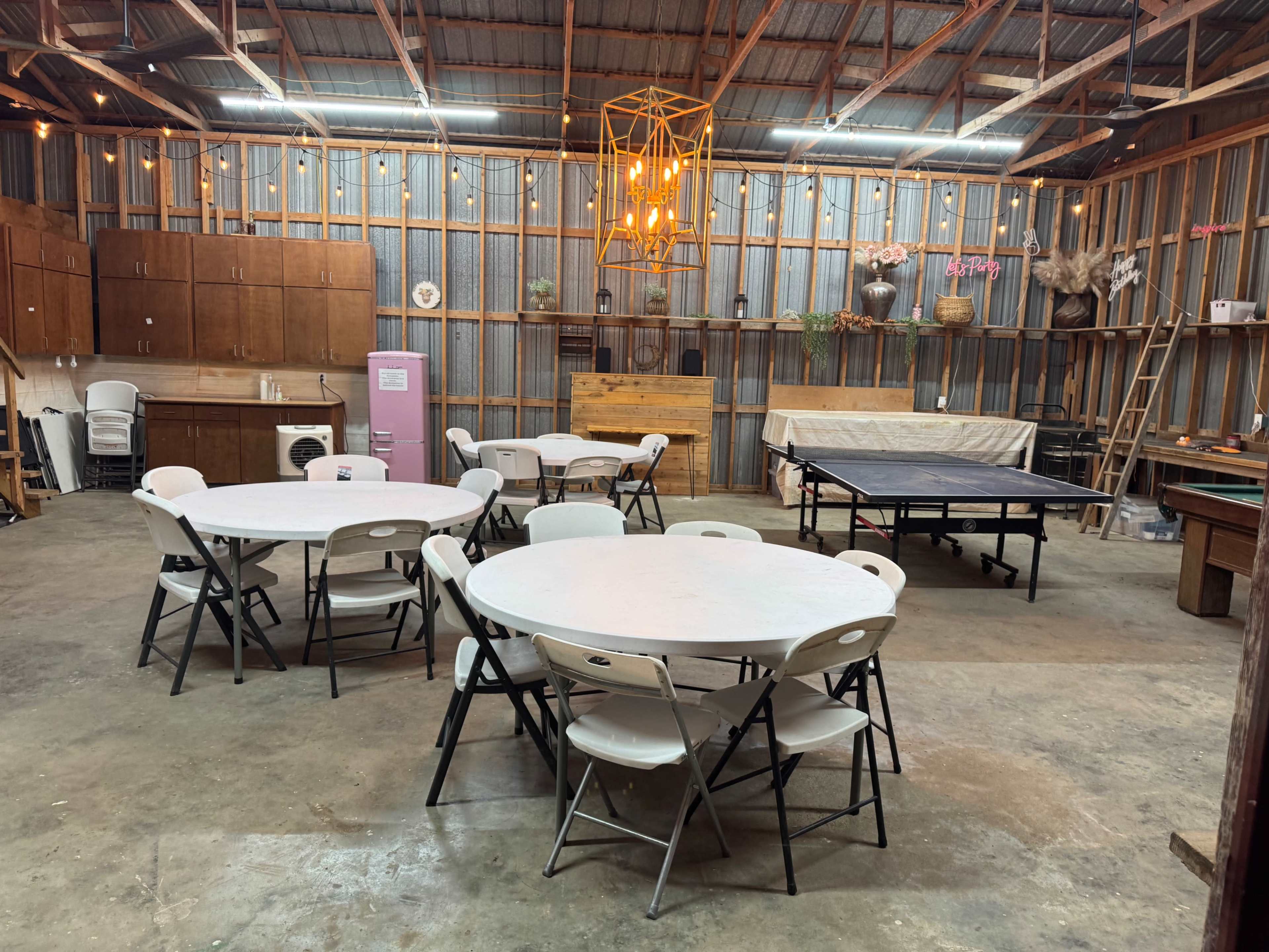 The image shows a spacious indoor area with circular tables and chairs arranged under a light fixture, alongside a ping pong table and storage units against the walls.