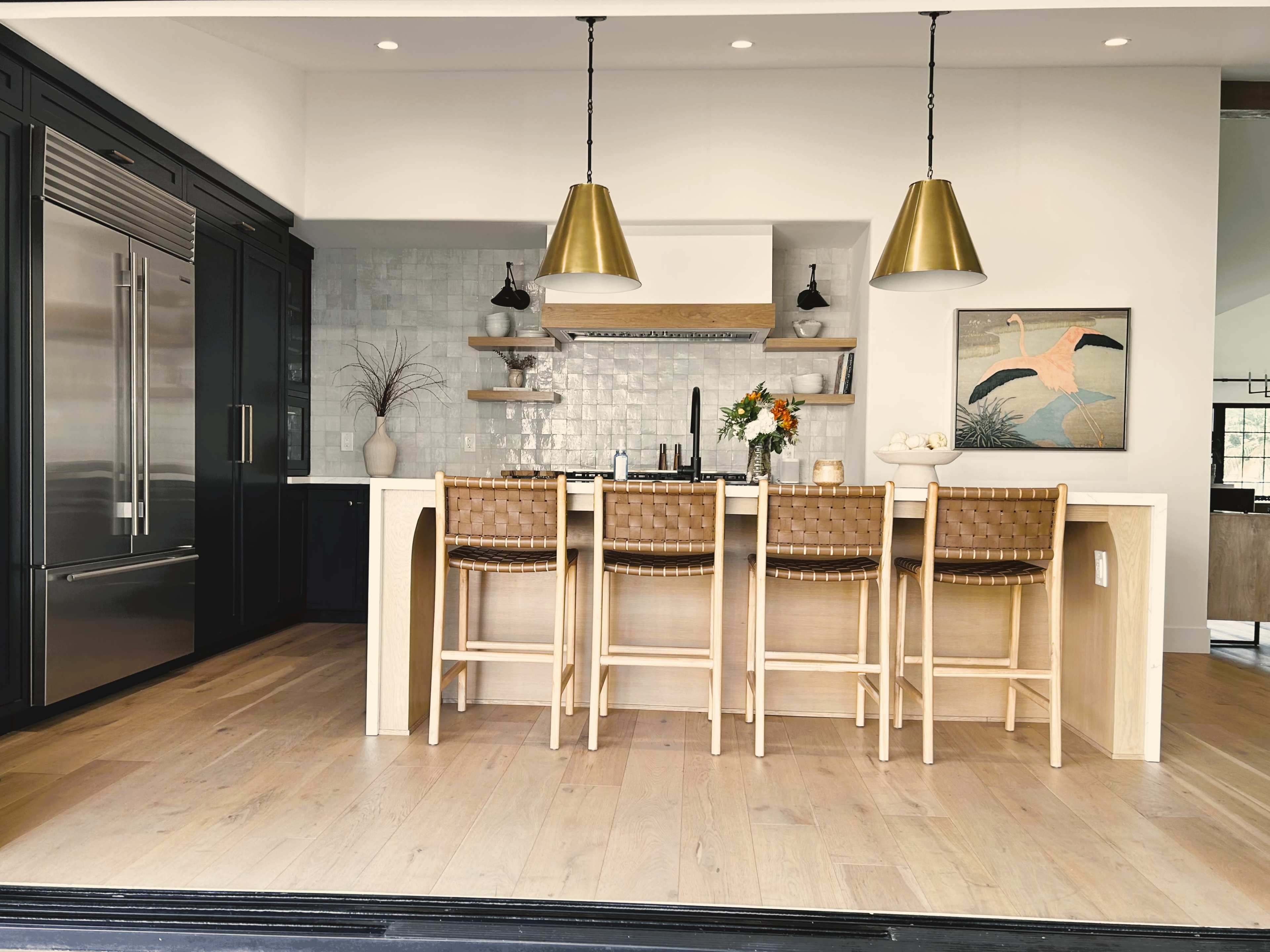 A modern kitchen featuring a light wood island with four bar stools, stainless steel appliances, and pendant lights above.