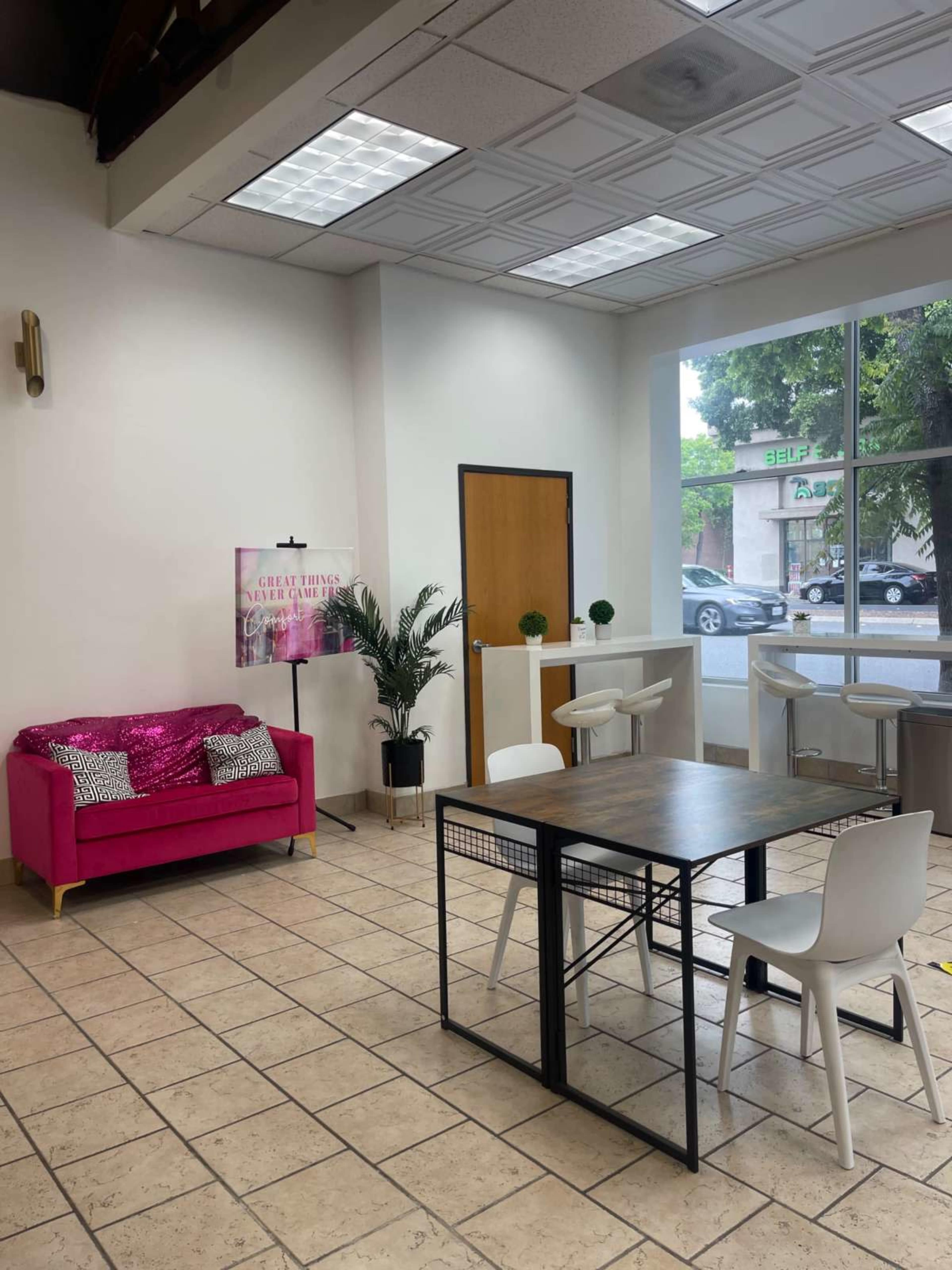 A bright interior space featuring a pink couch, a dining table with white chairs, and a decorative plant, with large windows providing natural light.