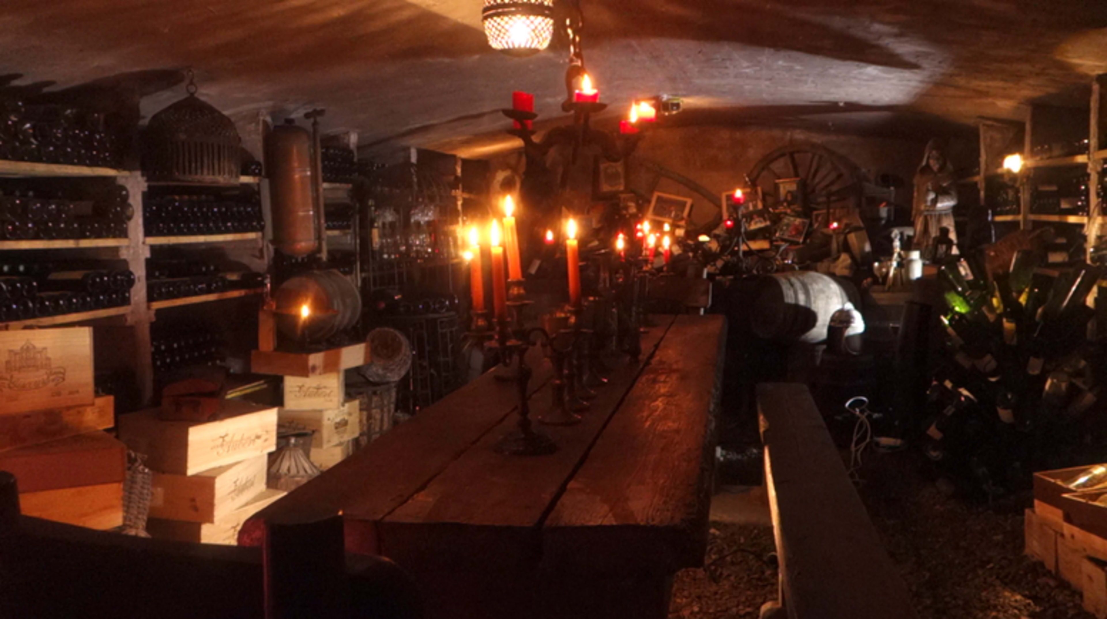 A dimly lit wine cellar filled with wooden storage boxes, bottles of wine, and candles on a long wooden table.