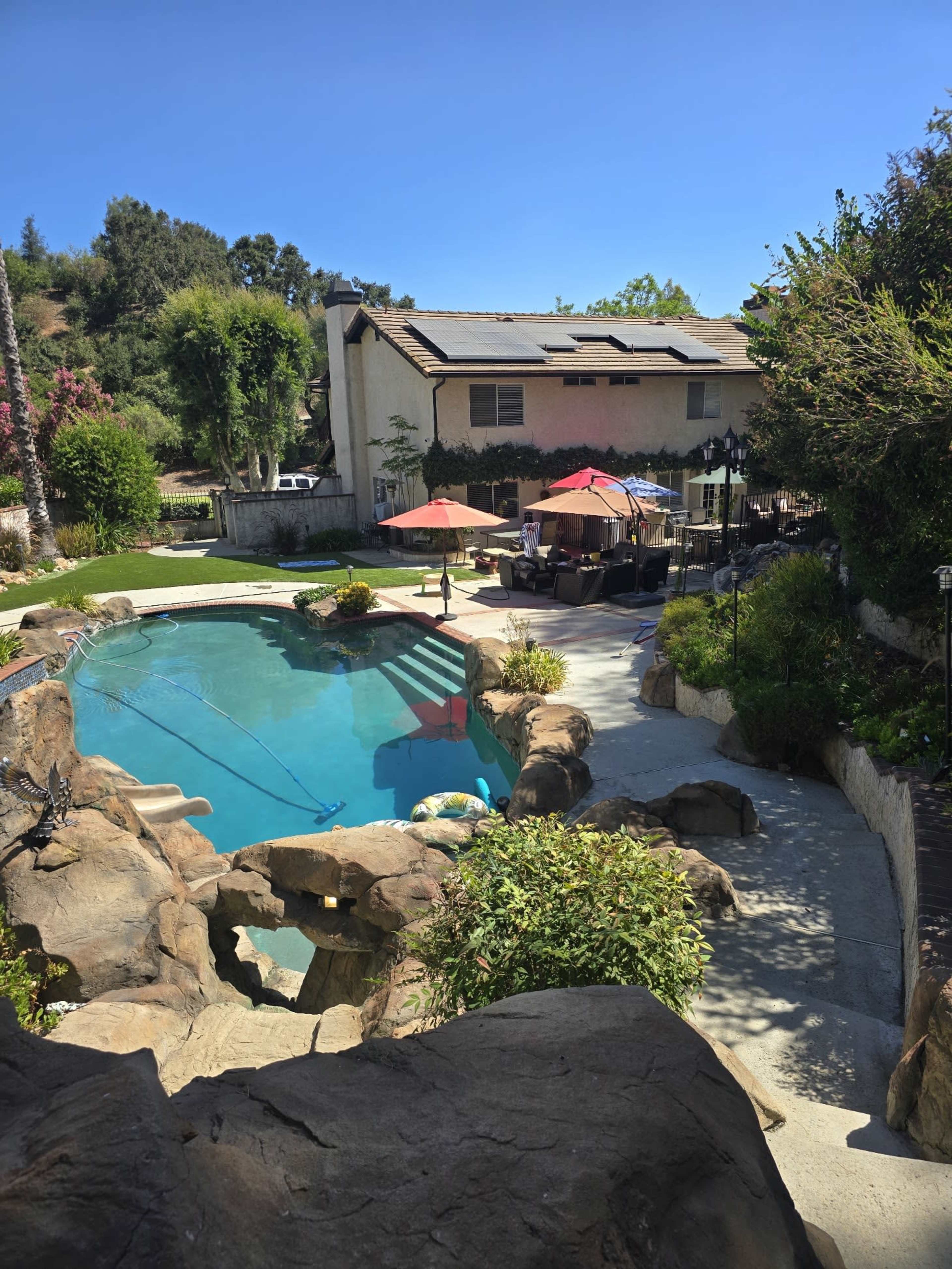 The image shows a residential backyard featuring a swimming pool surrounded by natural rock formations, lounge chairs, and patio umbrellas.