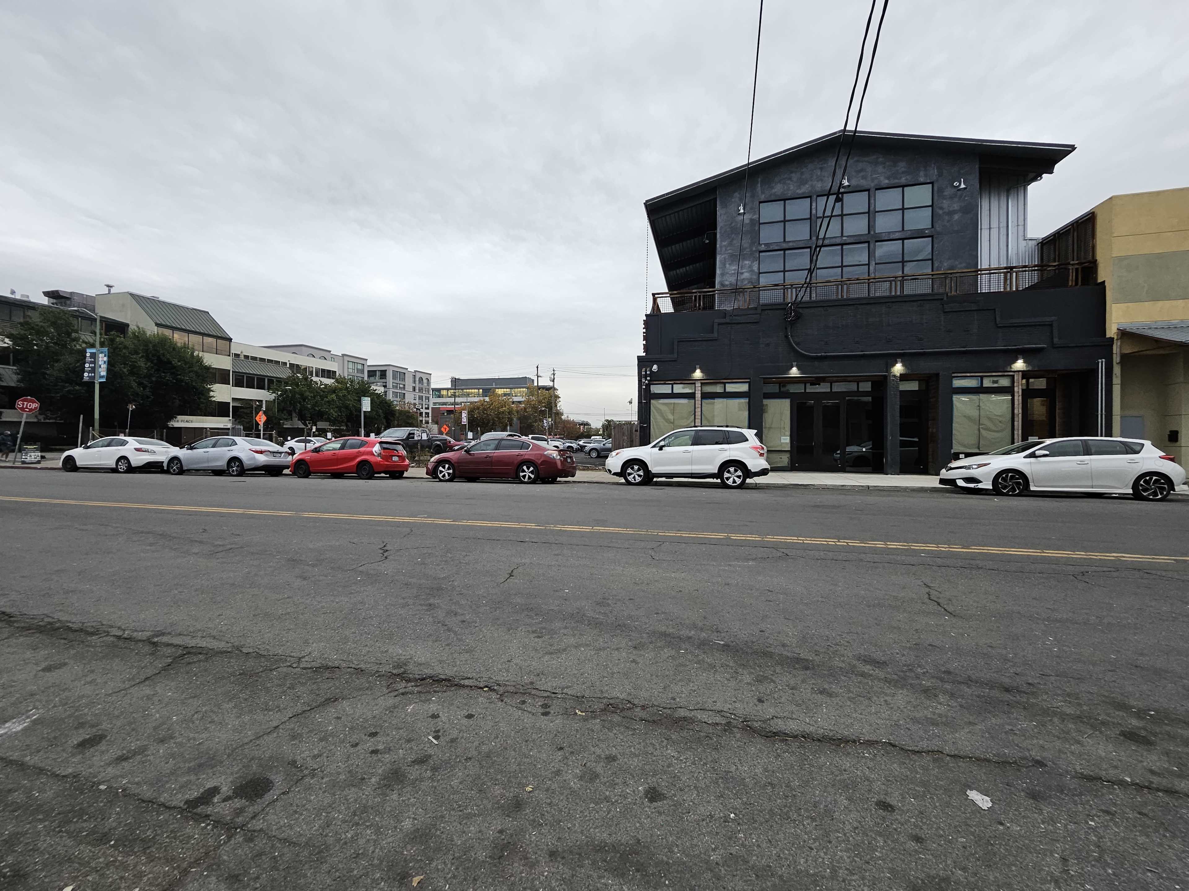 A line of parked cars is aligned along a street beside a two-story building with large windows and a flat roof.
