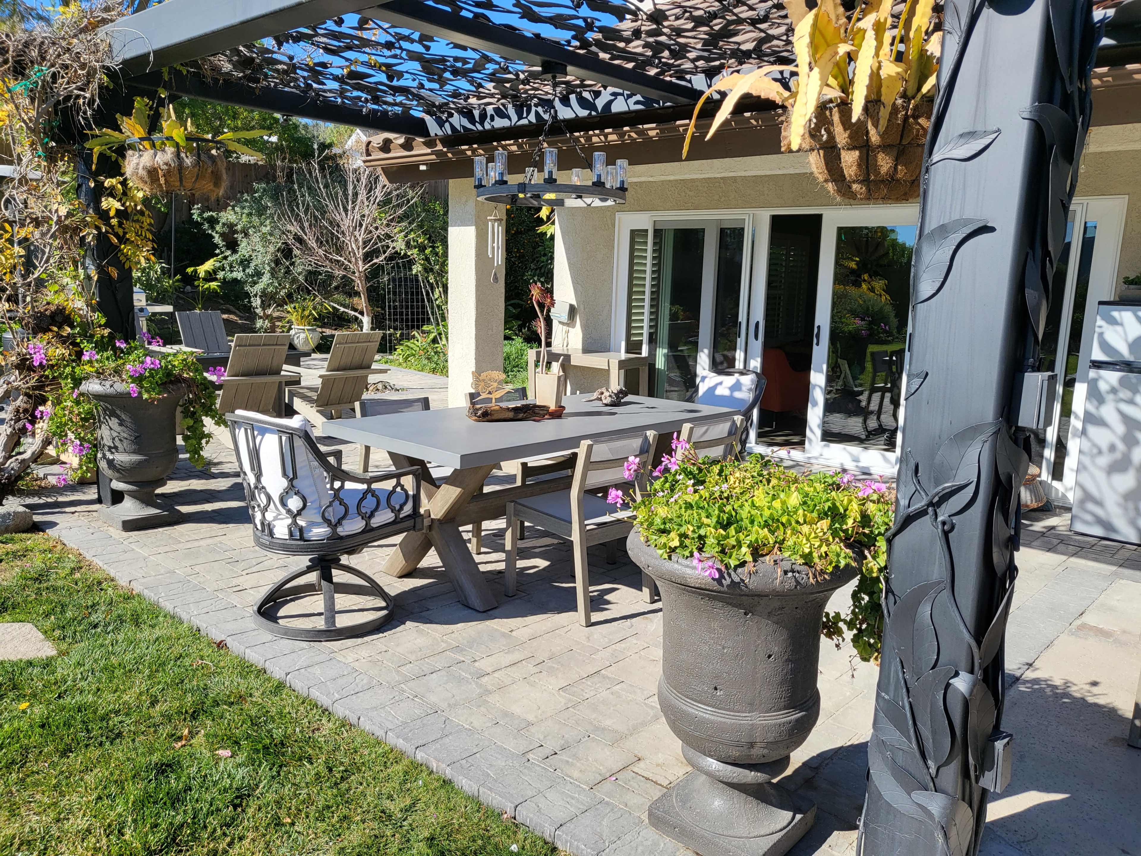 The image shows an outdoor dining area with a rectangular table surrounded by chairs, under a pergola adorned with plants and flowers, leading to a house in the background.