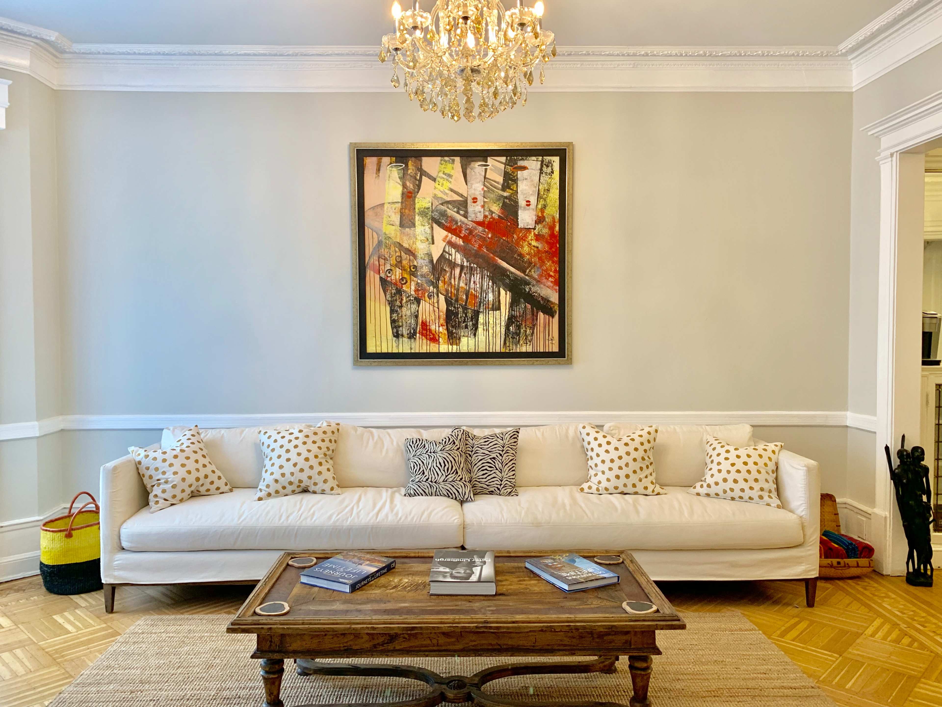 A white sofa with patterned pillows faces a wooden coffee table in a living room featuring a large abstract painting and a chandelier above.