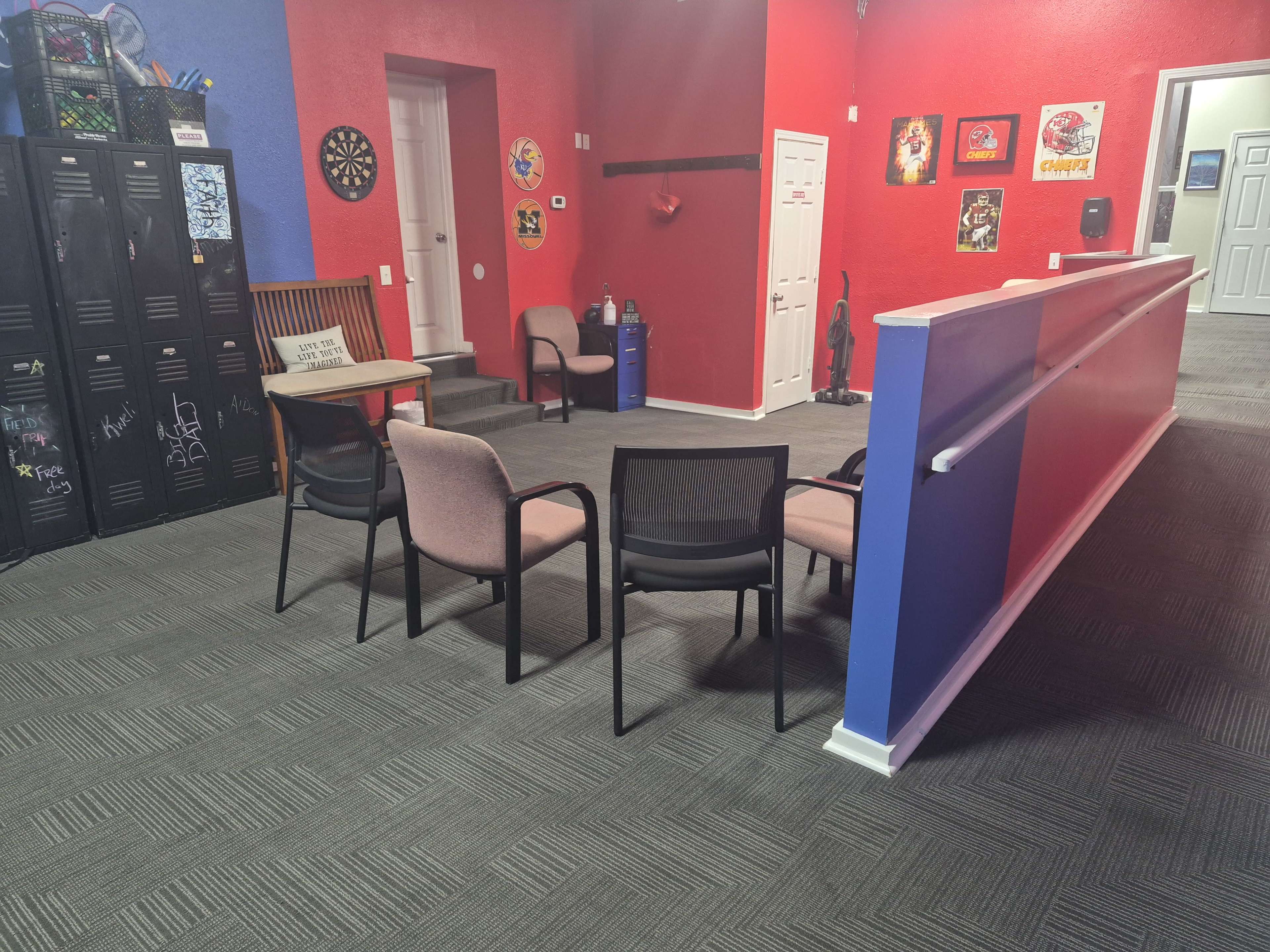 The image shows a room with red and blue walls, featuring several chairs arranged around a small table, lockers on one side, and framed artwork hanging on the walls.