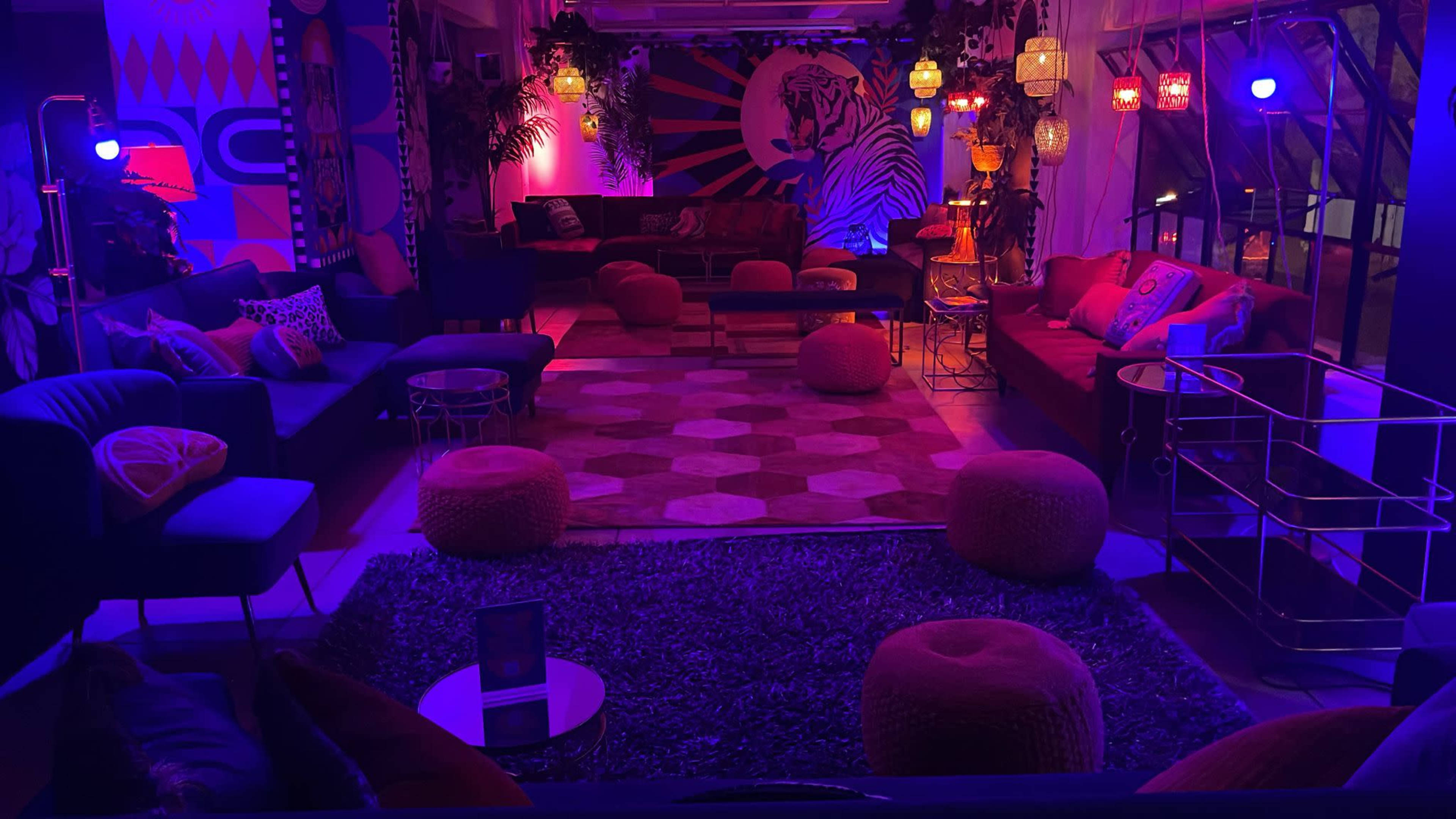 The scene features a vibrant lounge area with colorful seating, patterned rugs, decorative lighting, and plants.