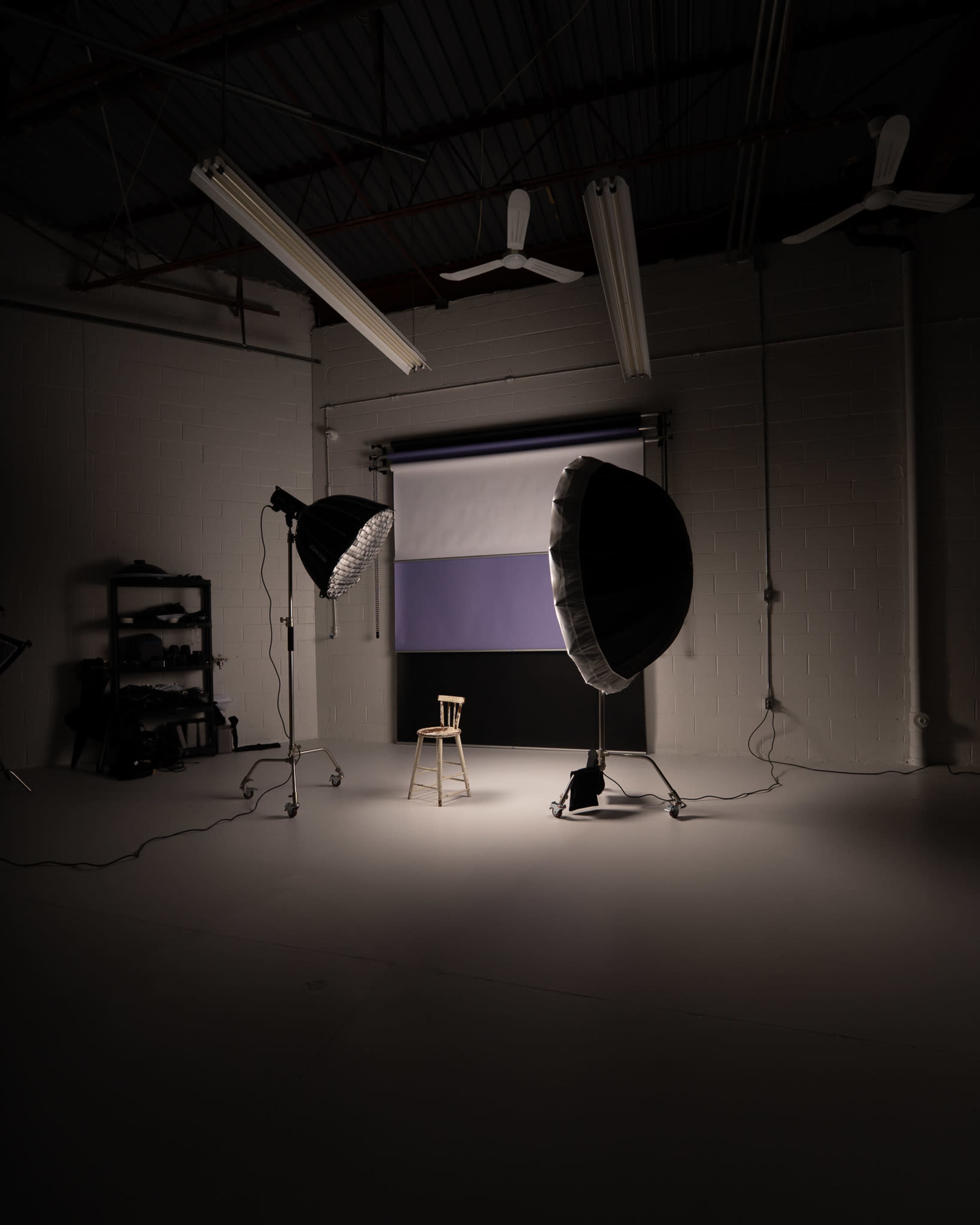 A single wooden chair is illuminated in a photography studio surrounded by two large light sources and a backdrop of purple and white.