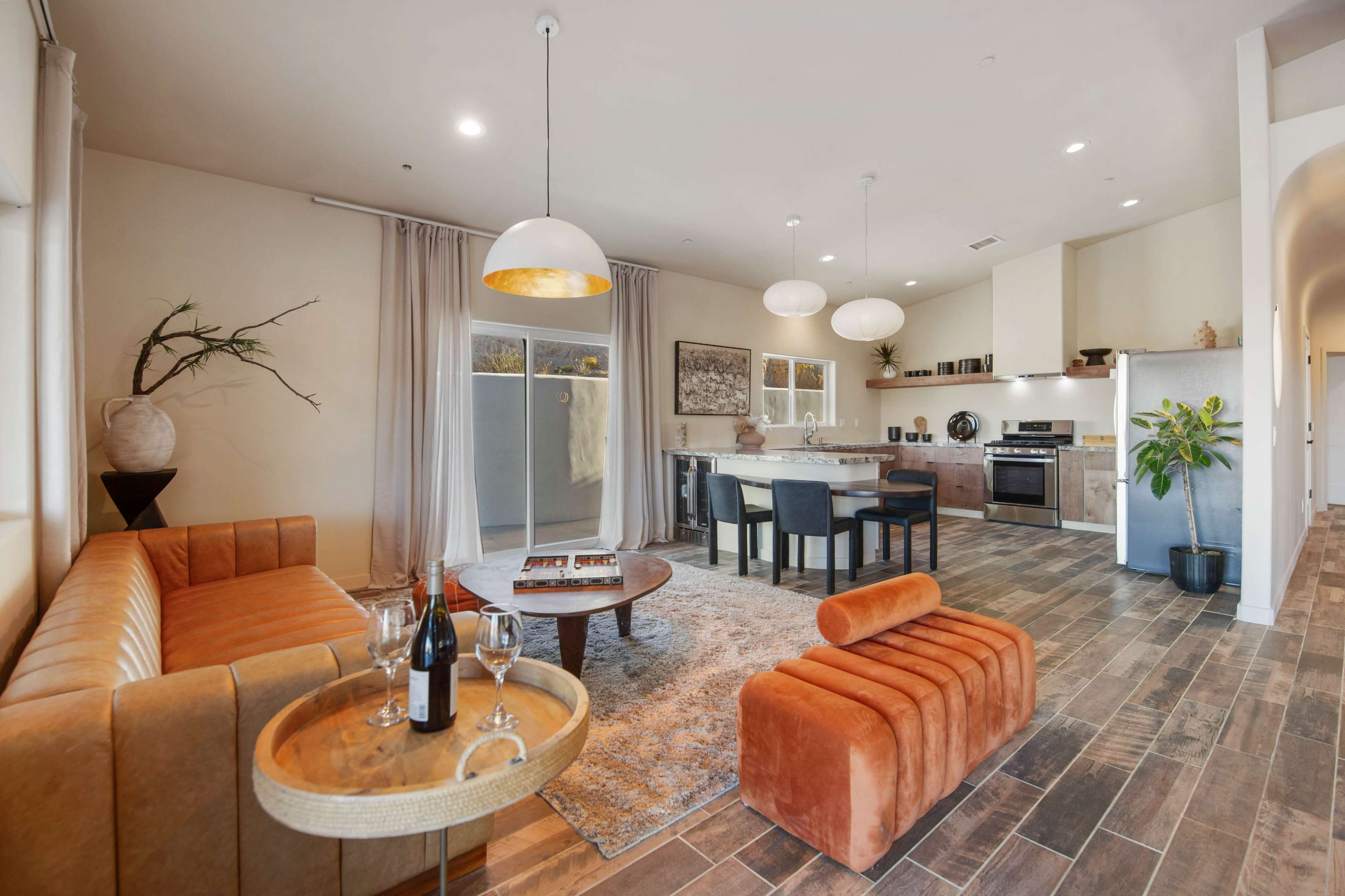 A modern living room features an orange sofa, a round coffee table with wine, and a kitchen area with a large island and appliances.