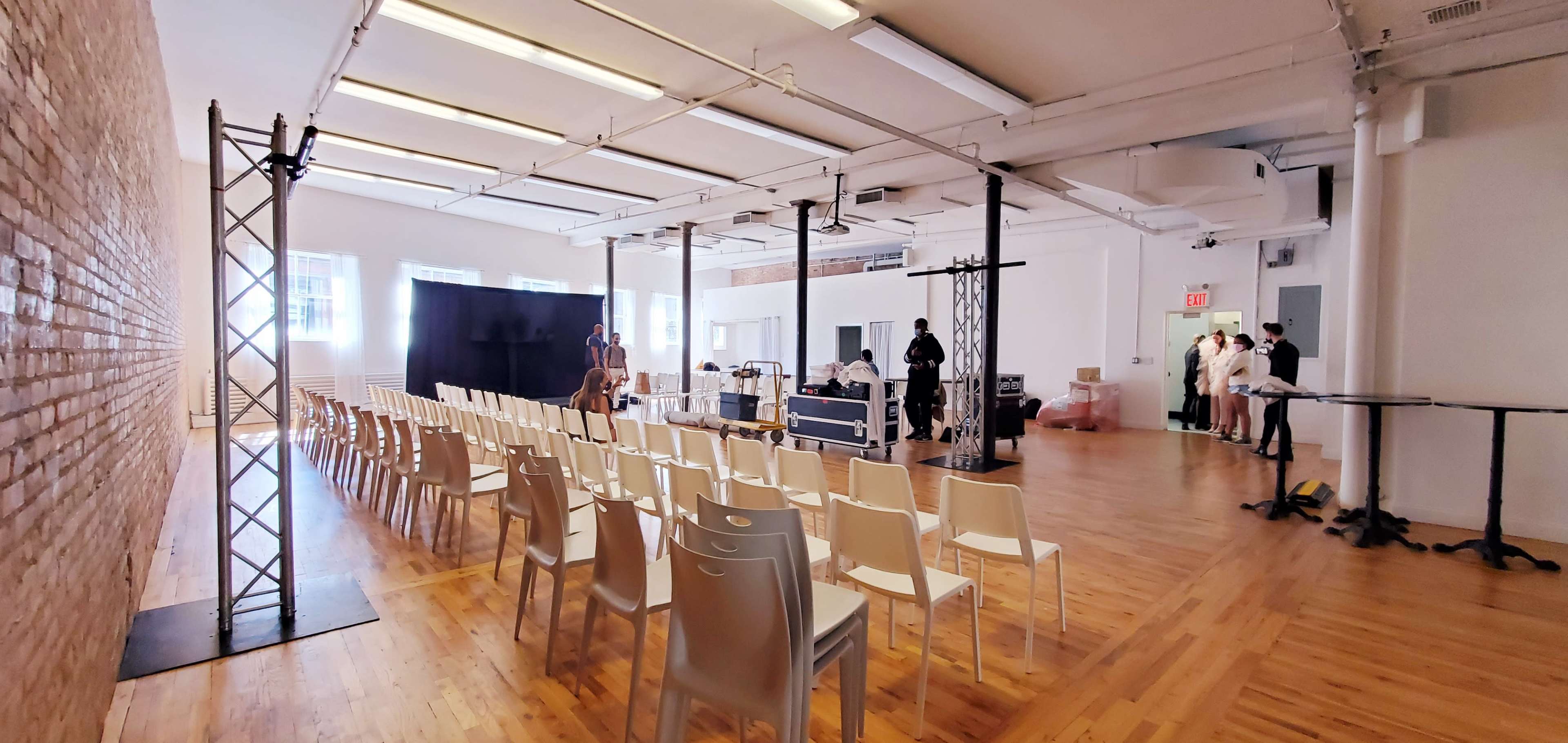 The image shows a spacious room with rows of empty chairs arranged facing a black backdrop, while several people prepare equipment and a stage setup.