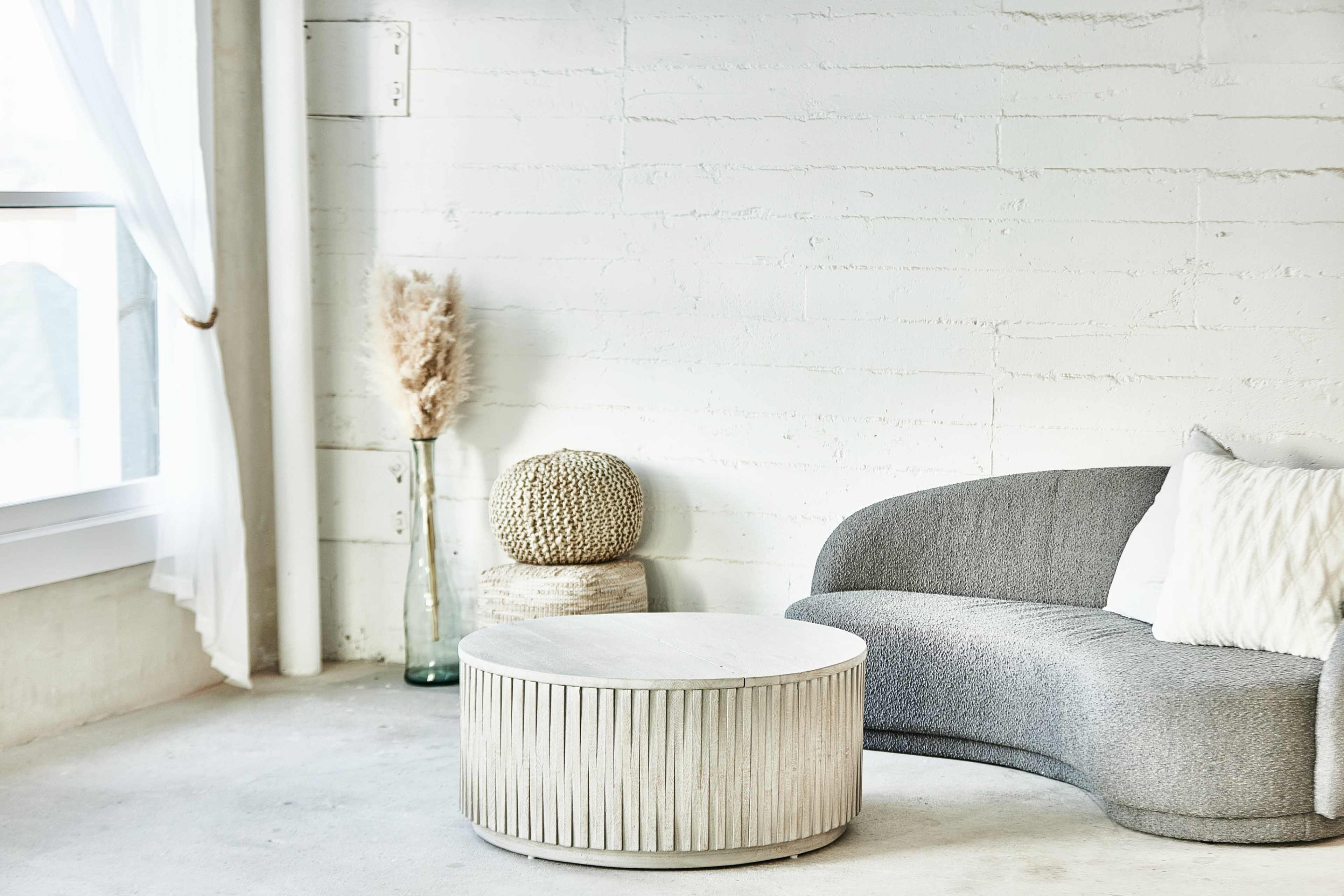 A minimalist living area features a curved gray sofa, a round wooden coffee table with a textured design, and decorative elements including a woven basket and a glass vase holding pampas grass.