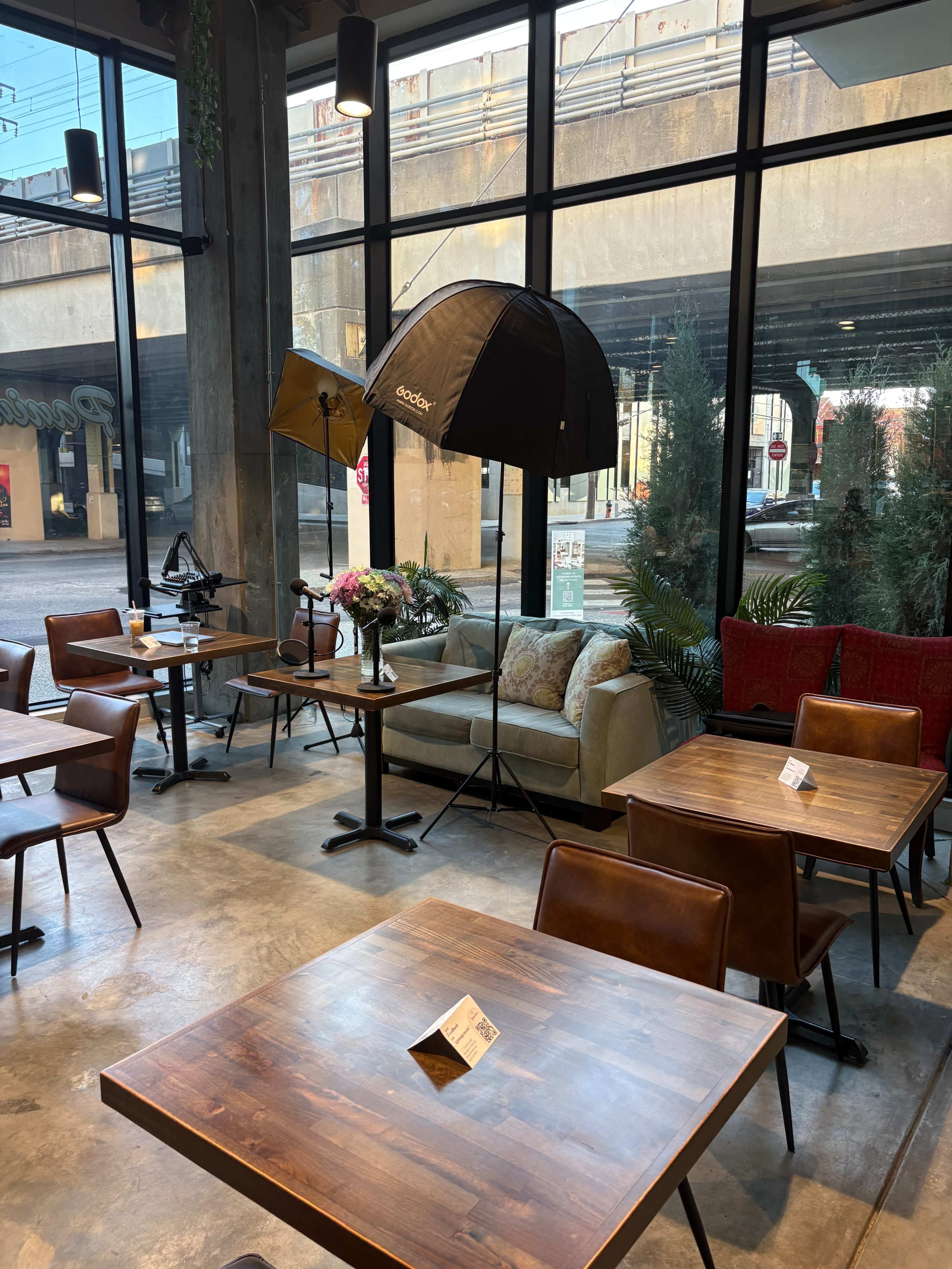 The image shows a café interior with wooden tables and chairs, a sofa, and photography equipment including softboxes near a large window.