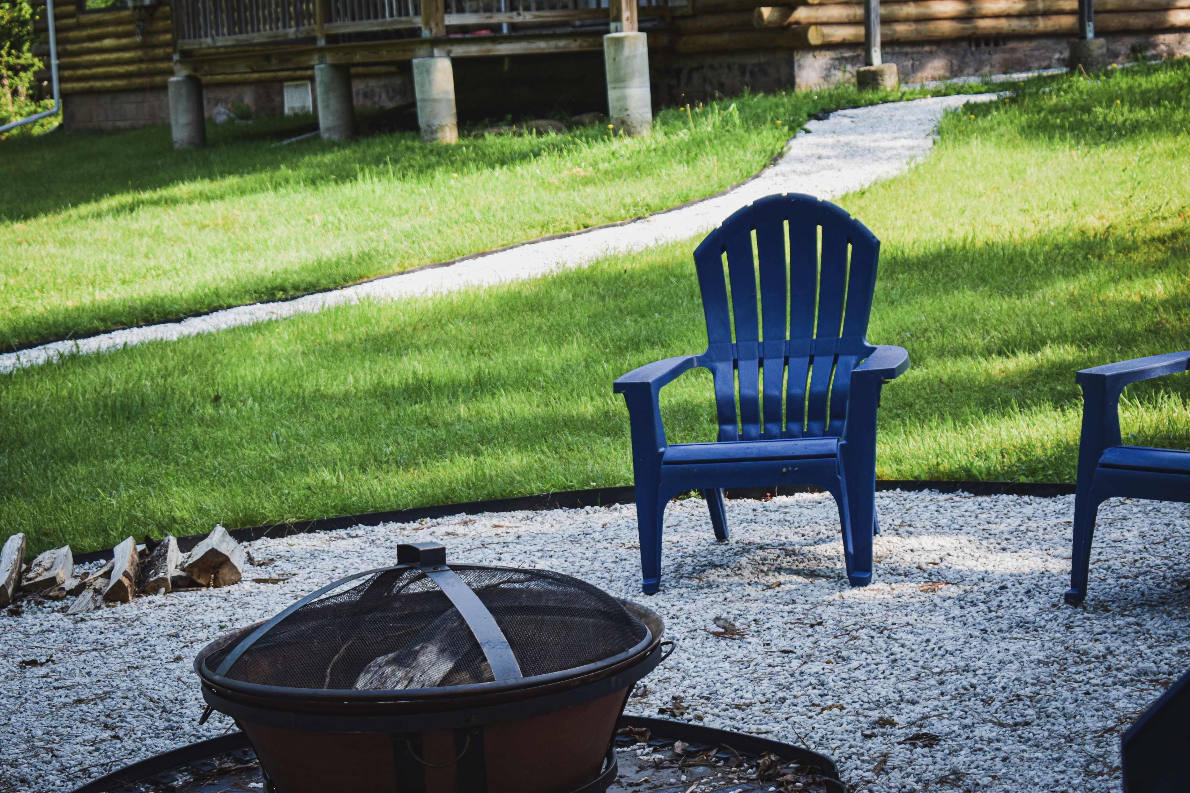 The image shows a fire pit surrounded by gravel, with two blue adirondack chairs placed on the grassy lawn near a winding path.