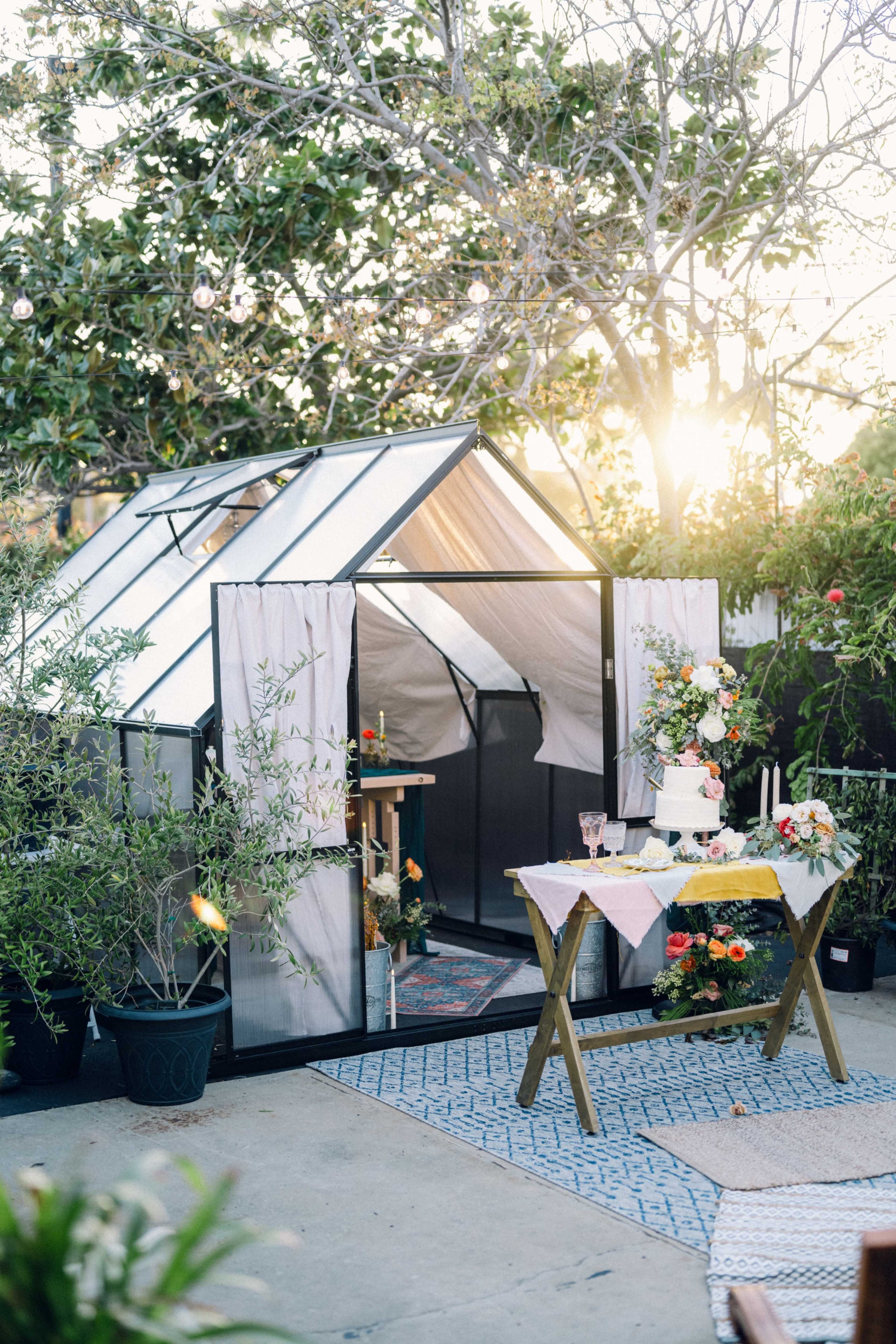 A greenhouse stands in a sunlit garden beside a decorated table with floral arrangements.