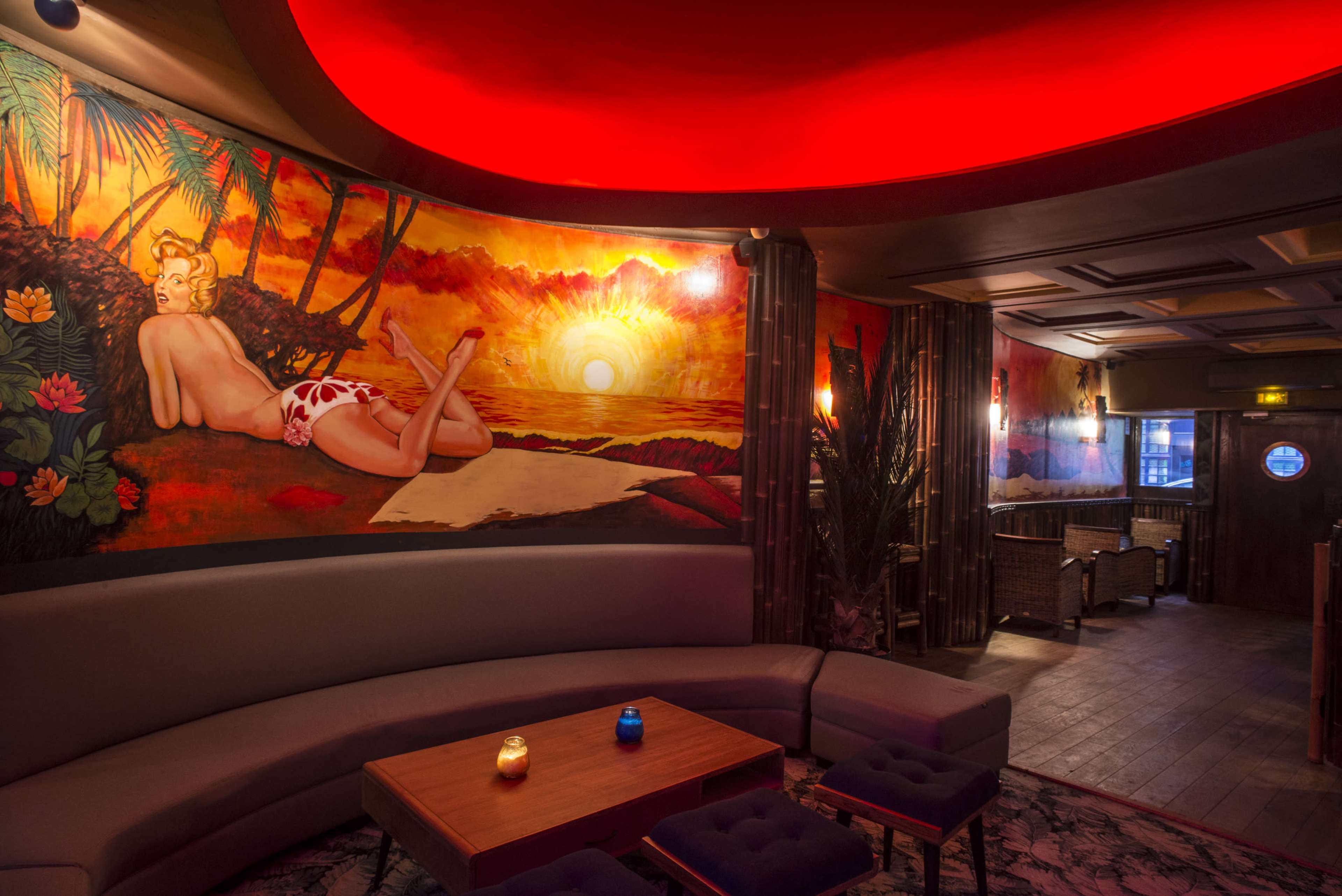 The image shows a lounge with a curved sofa, a wooden table, and vibrant wall art depicting a sunset and a reclining figure.