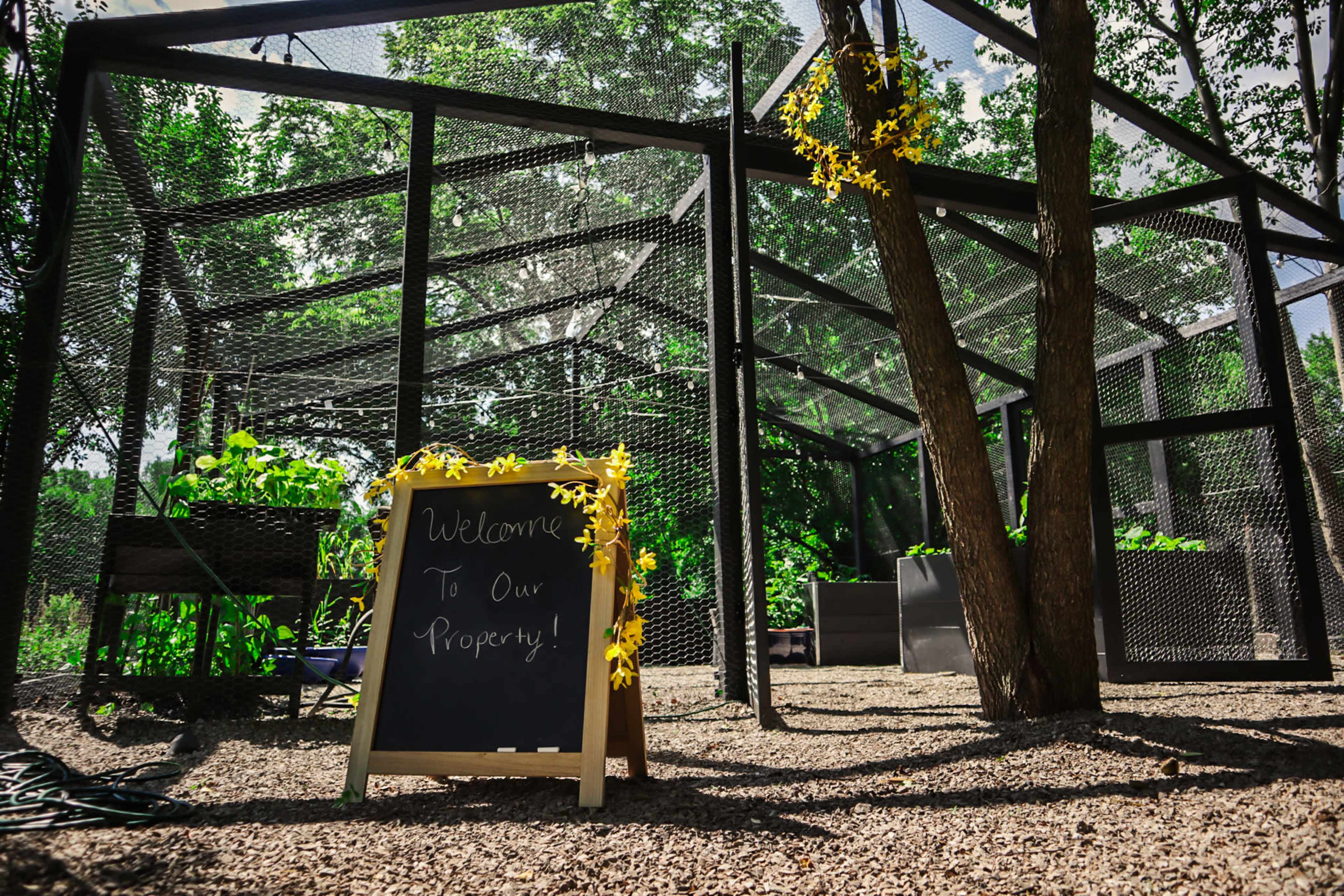 A garden enclosure with a chalkboard sign that reads, "Welcome To Our Property!" is surrounded by greenery and trees.