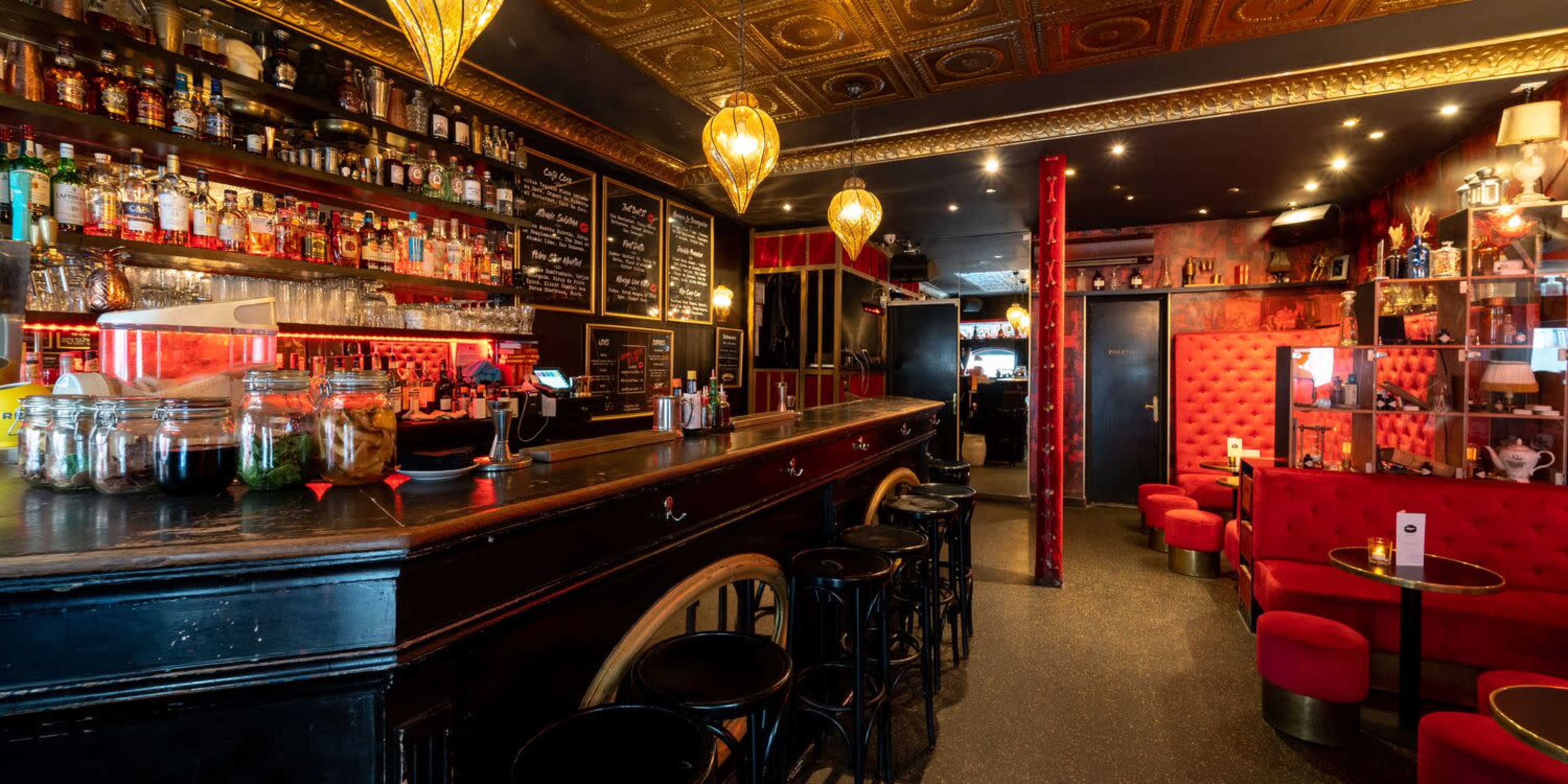 A stylish bar interior featuring a wooden bar counter, shelves stocked with various bottles, red upholstered seating, and decorative lighting.