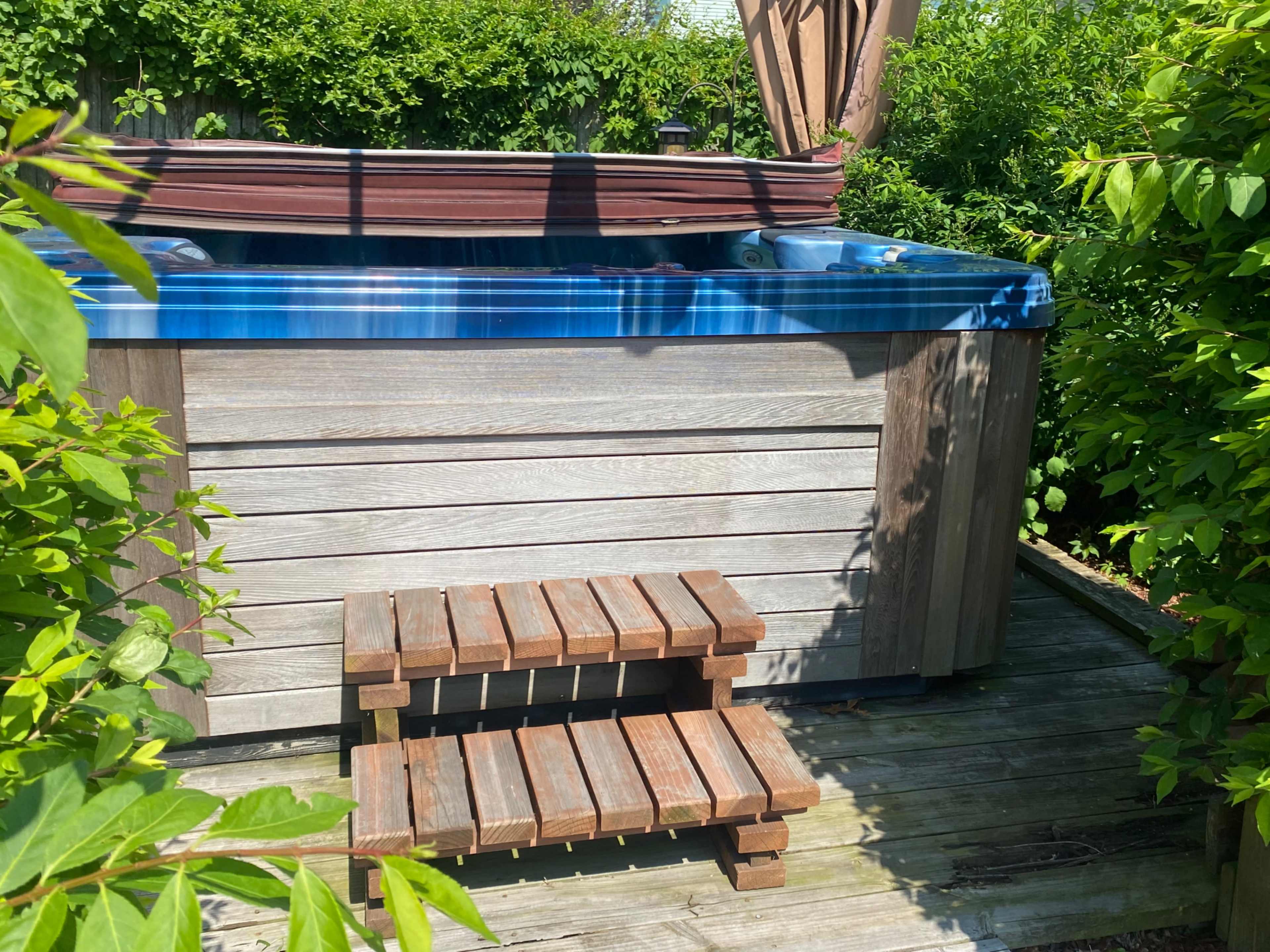 A wooden hot tub with a blue cover is situated on a deck surrounded by greenery, alongside a small wooden step stool.