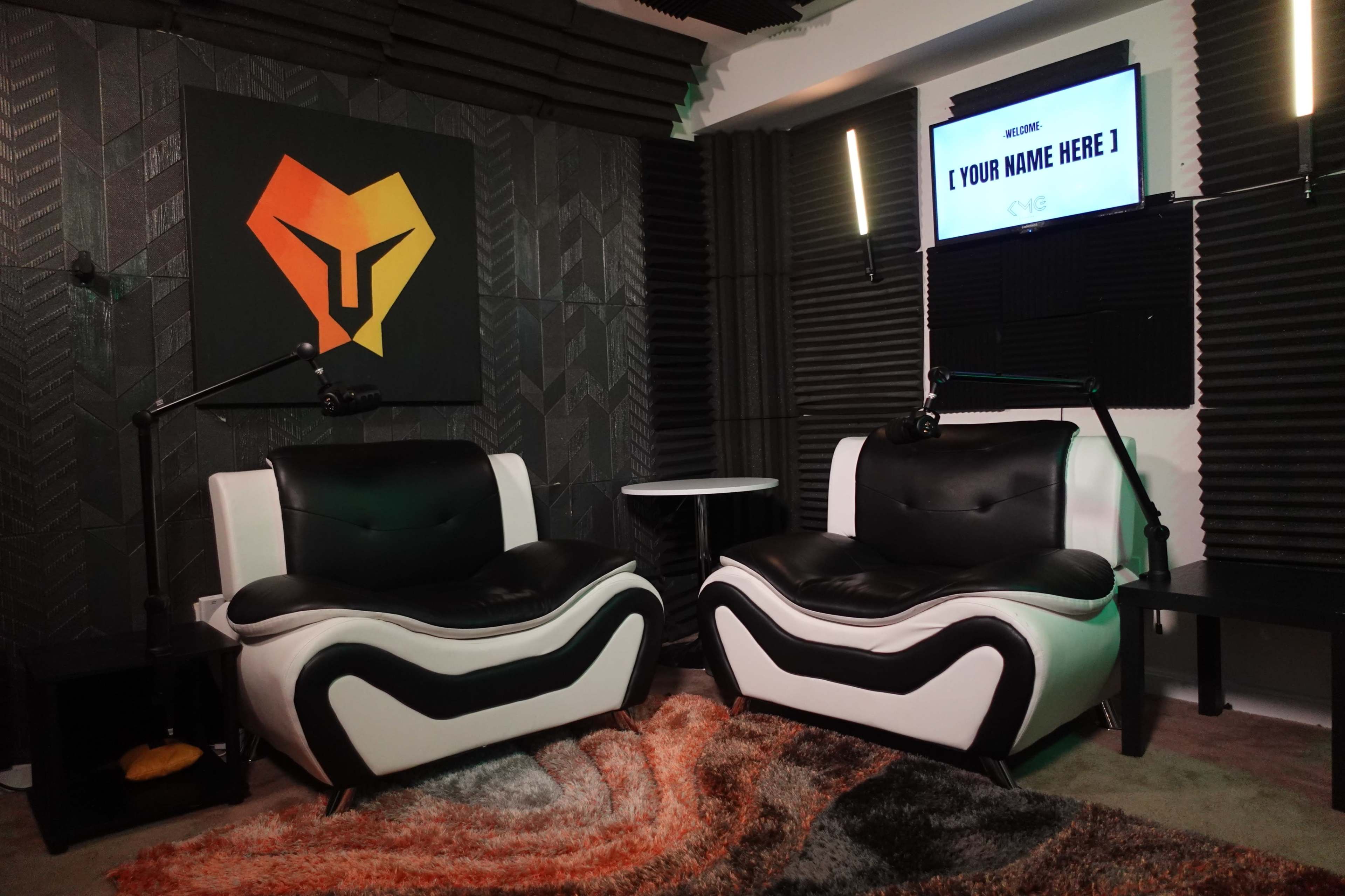The image shows a modern podcast studio featuring two black and white armchairs, a wall-mounted screen with a customizable title, and acoustic paneling behind the chairs.