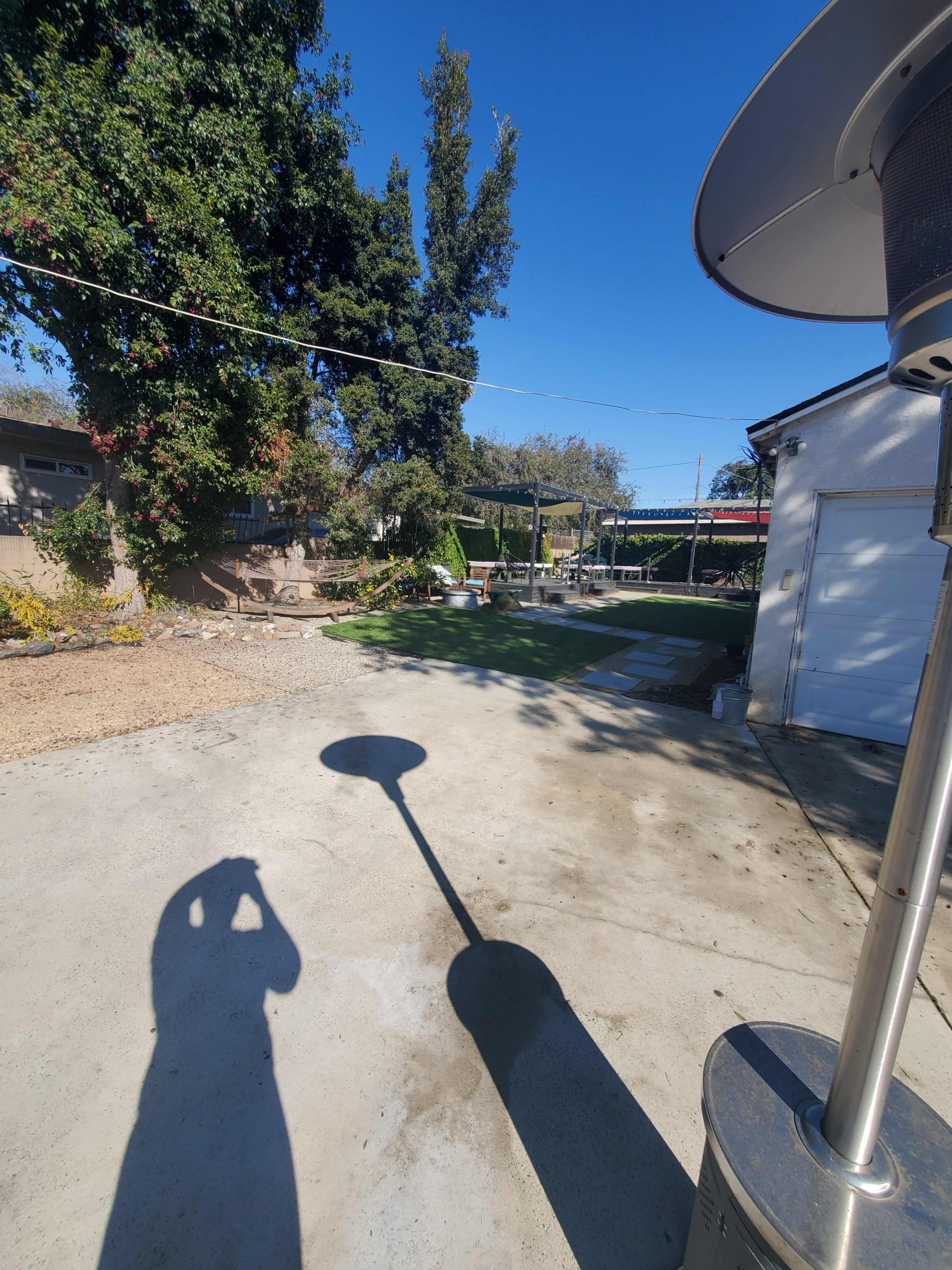 A backyard with a concrete surface, a patio heater casting a shadow, and various plants and structures in the background.