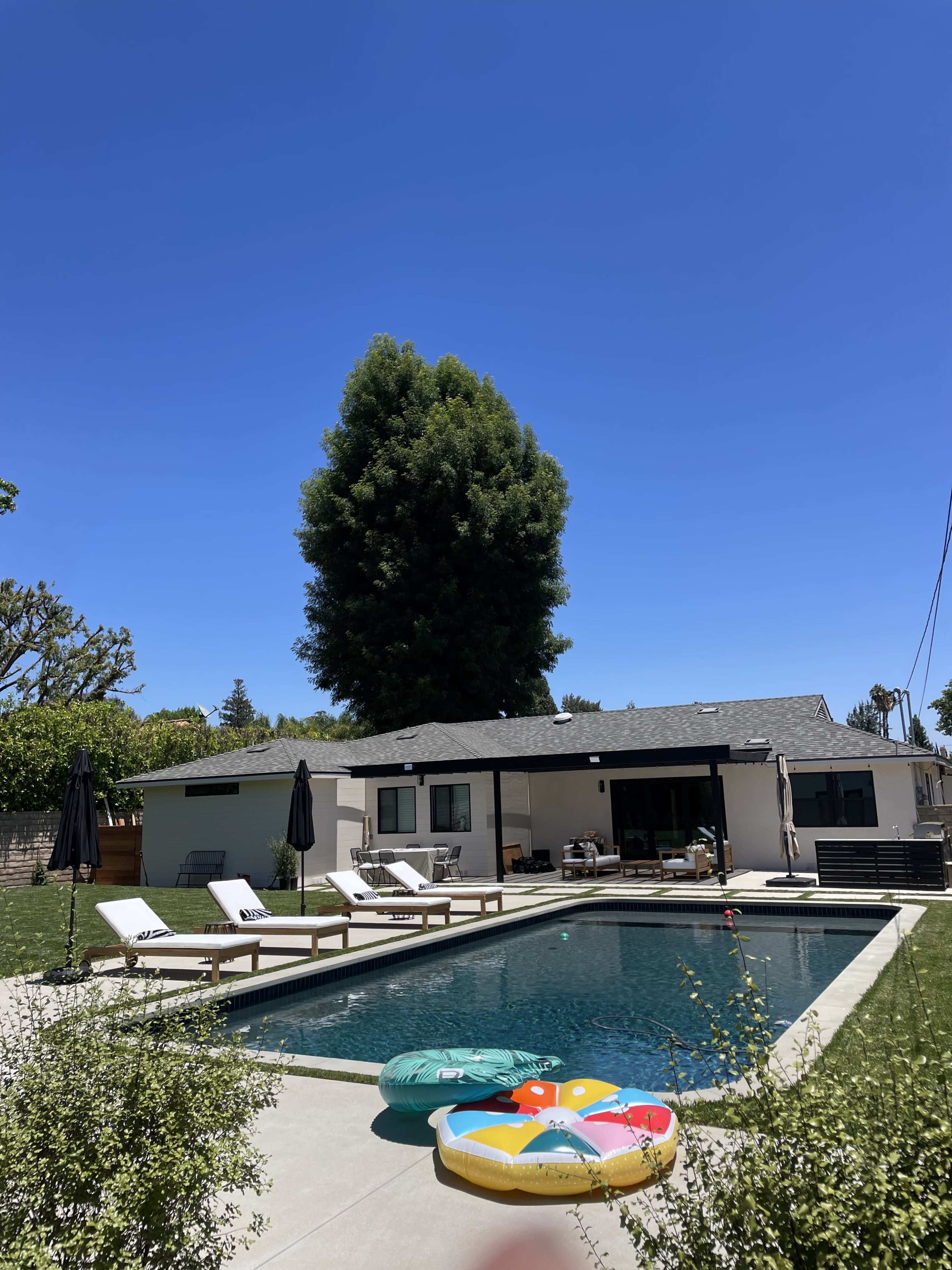 A backyard pool area features lounge chairs, a colorful pool float, and a modern house under a clear blue sky.