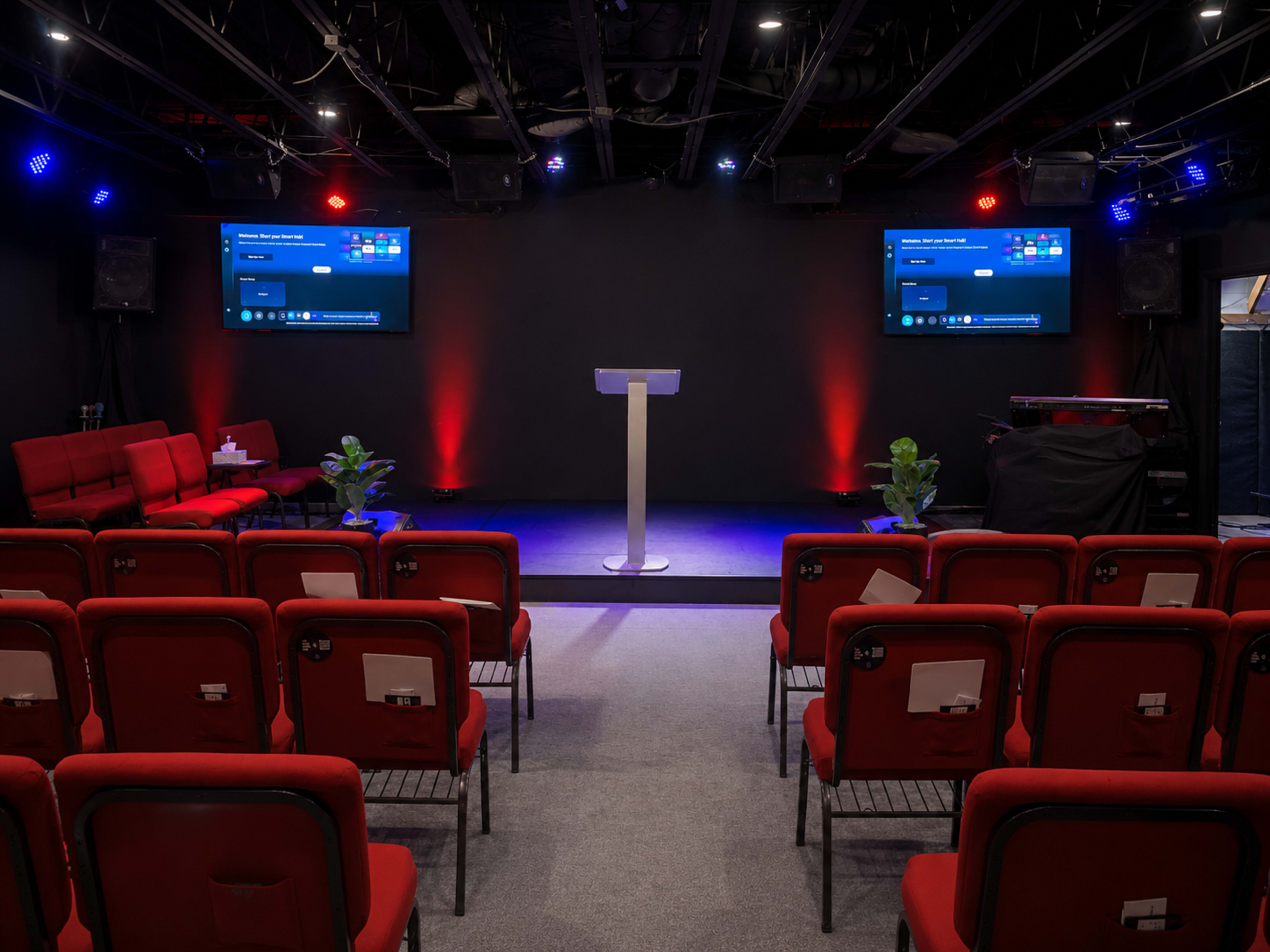 The image shows a small conference room or stage setup with red chairs arranged in rows, a central podium, and large screens mounted on the walls.
