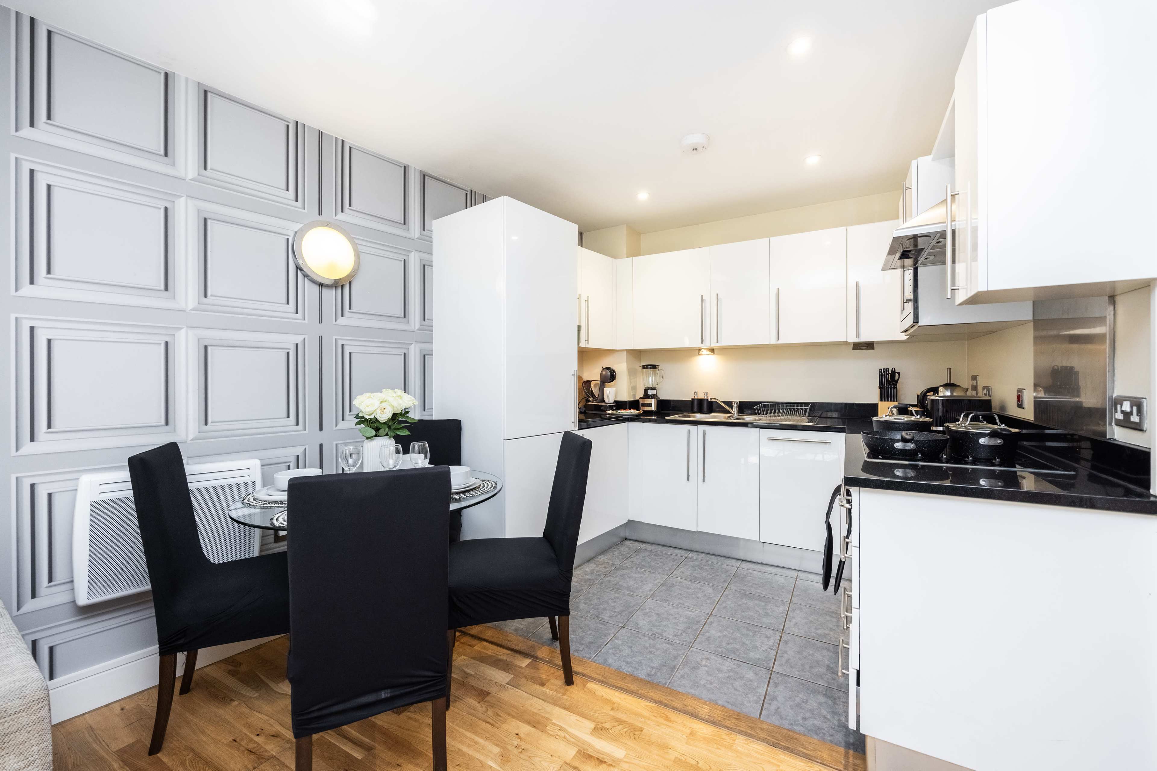 The image shows a modern kitchen with white cabinetry, a round dining table set for two, and gray paneling on one wall.