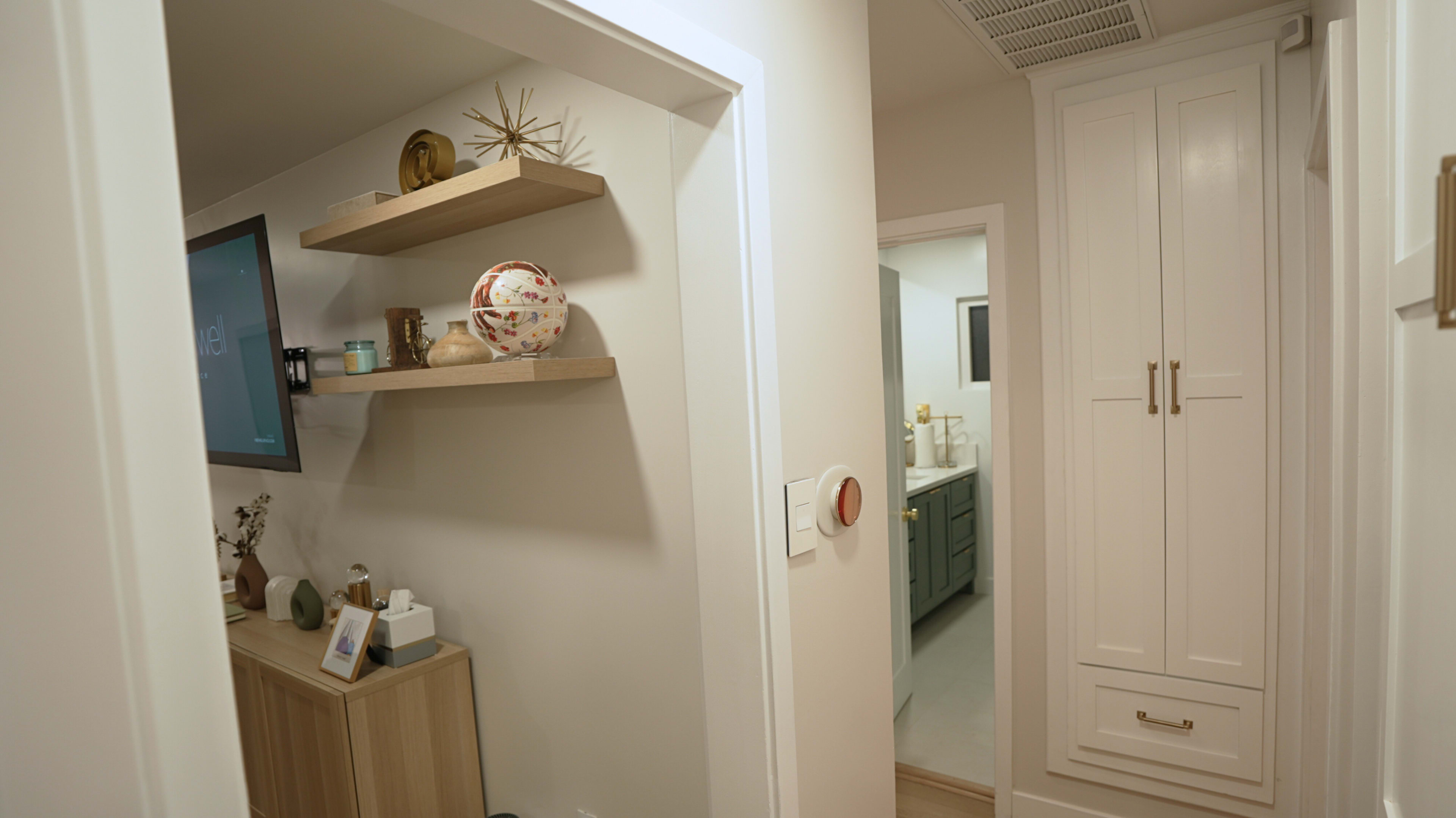 The image shows a modern hallway with a small wooden table and shelves displaying decorative items, leading to a bathroom or kitchen area visible in the background.