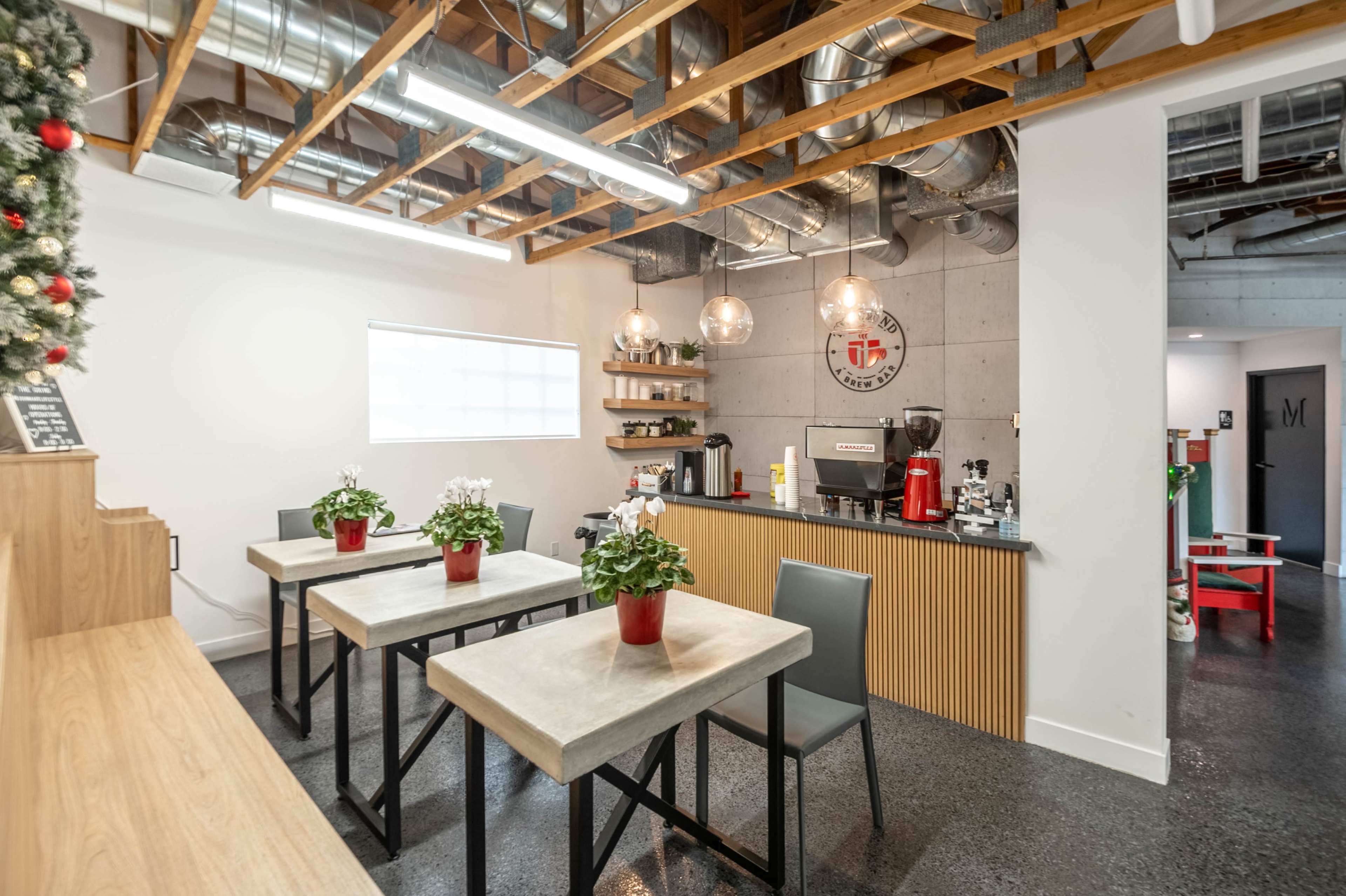The image shows a modern kitchen area with a few tables, each featuring a potted plant, and a countertop equipped with coffee-making appliances and decorative items.