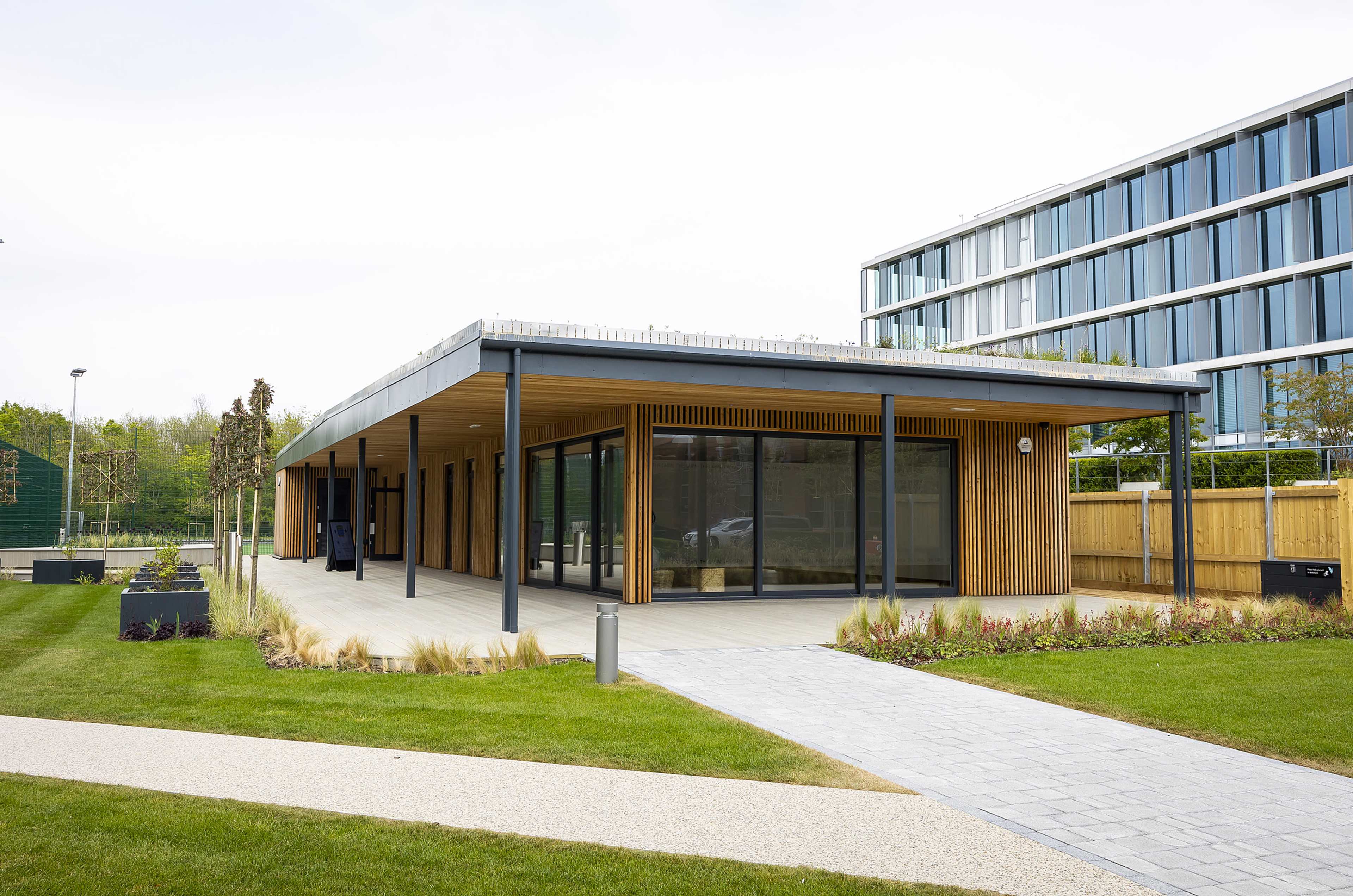 A modern building with a wooden facade and large glass windows, situated in a landscaped area featuring grass and paved pathways.