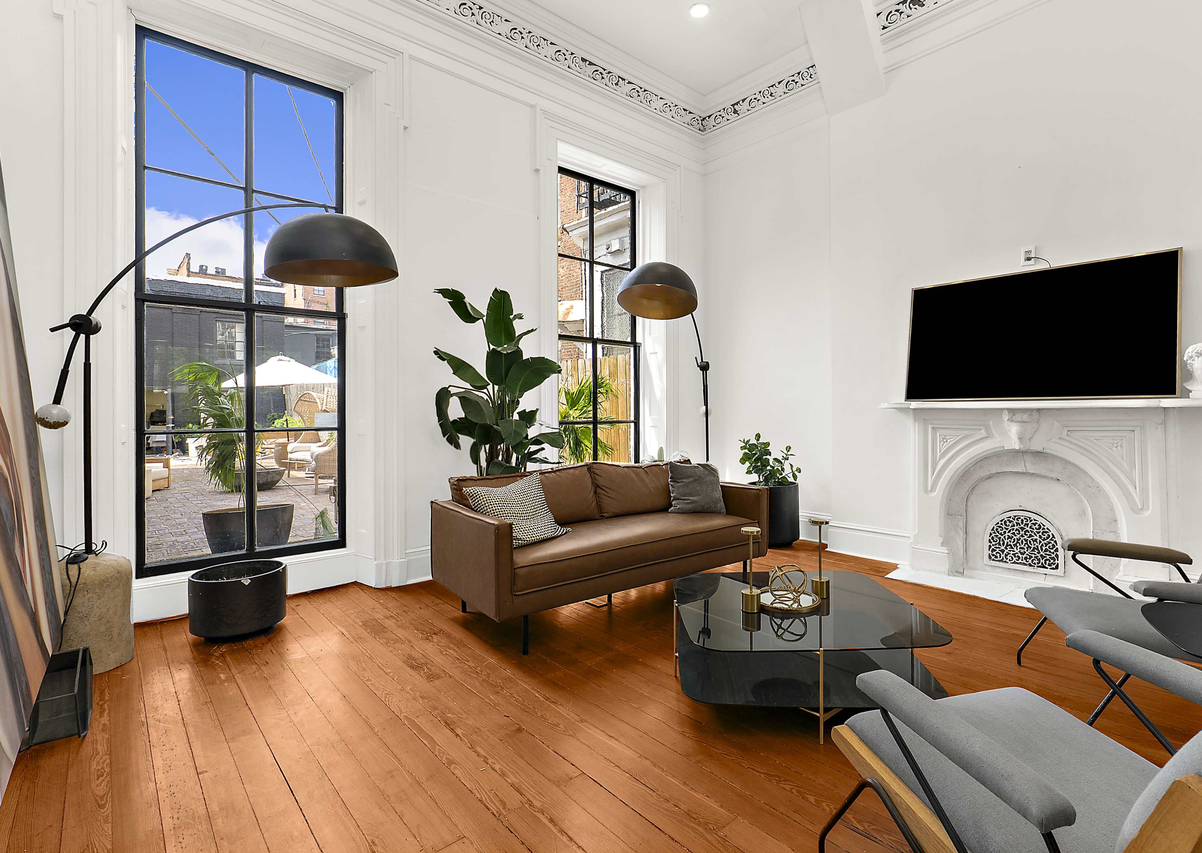 A modern living room features a brown leather sofa, two sleek chairs, a glass coffee table, and large windows that overlook a patio with outdoor furniture.