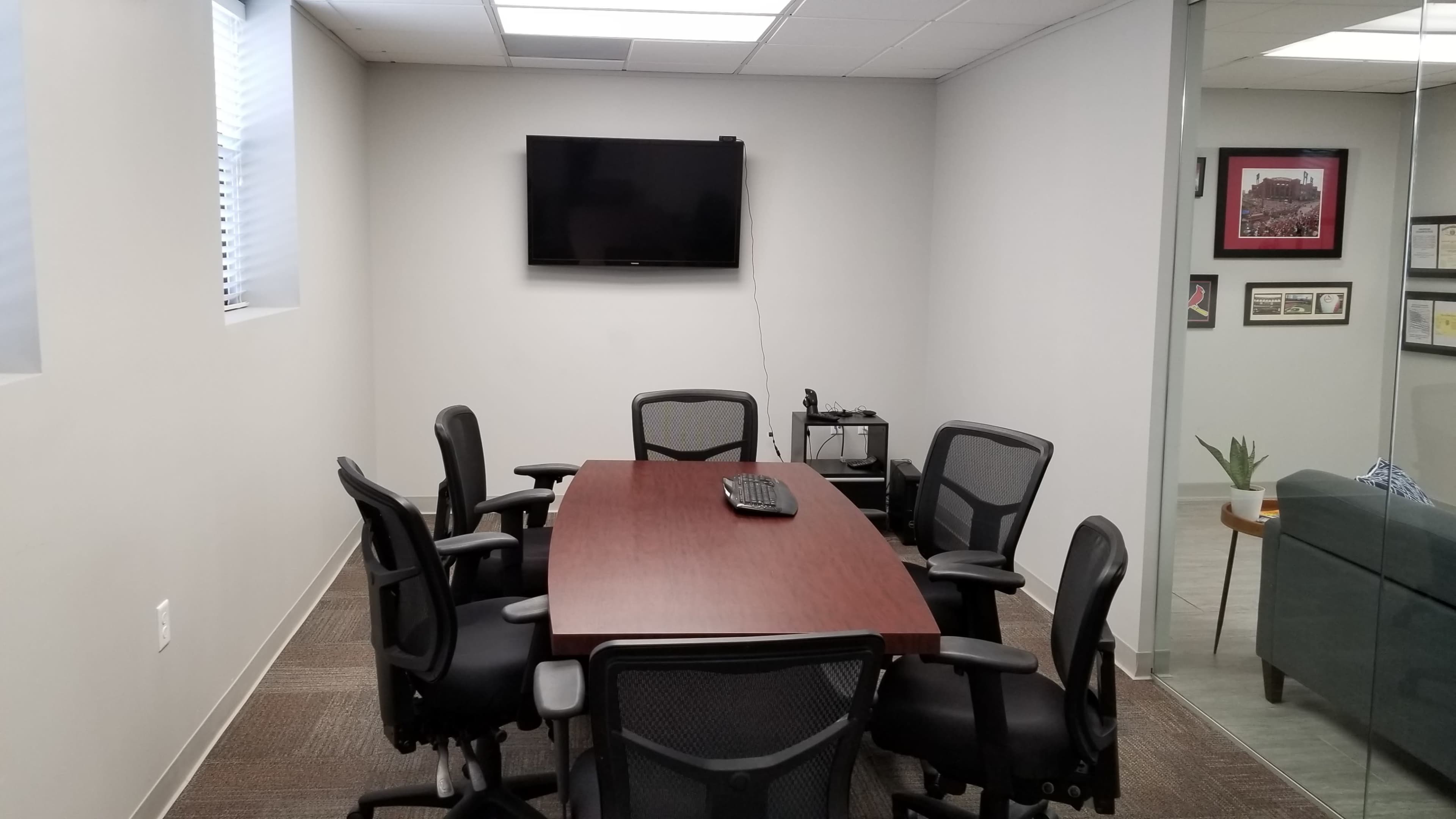 A meeting room features a rectangular wooden table surrounded by several black office chairs, with a wall-mounted TV and a glass wall displaying framed documents.