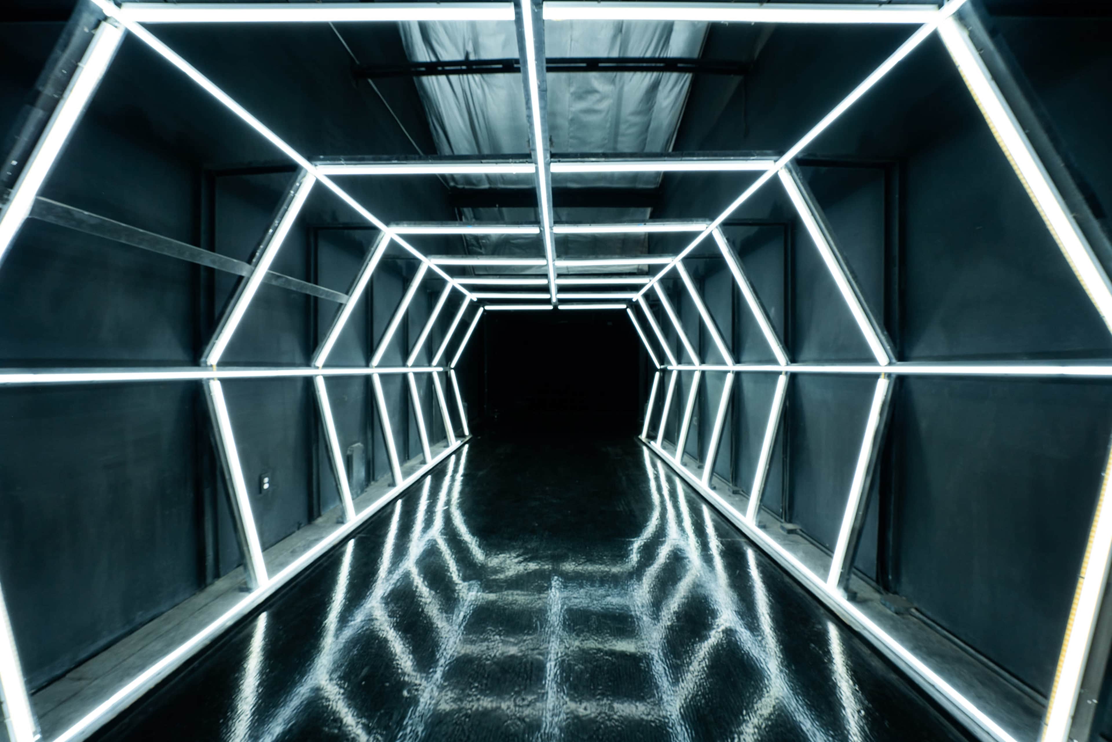 The image shows a dimly lit corridor with geometric, illuminated outlines created by bright, white neon lights.