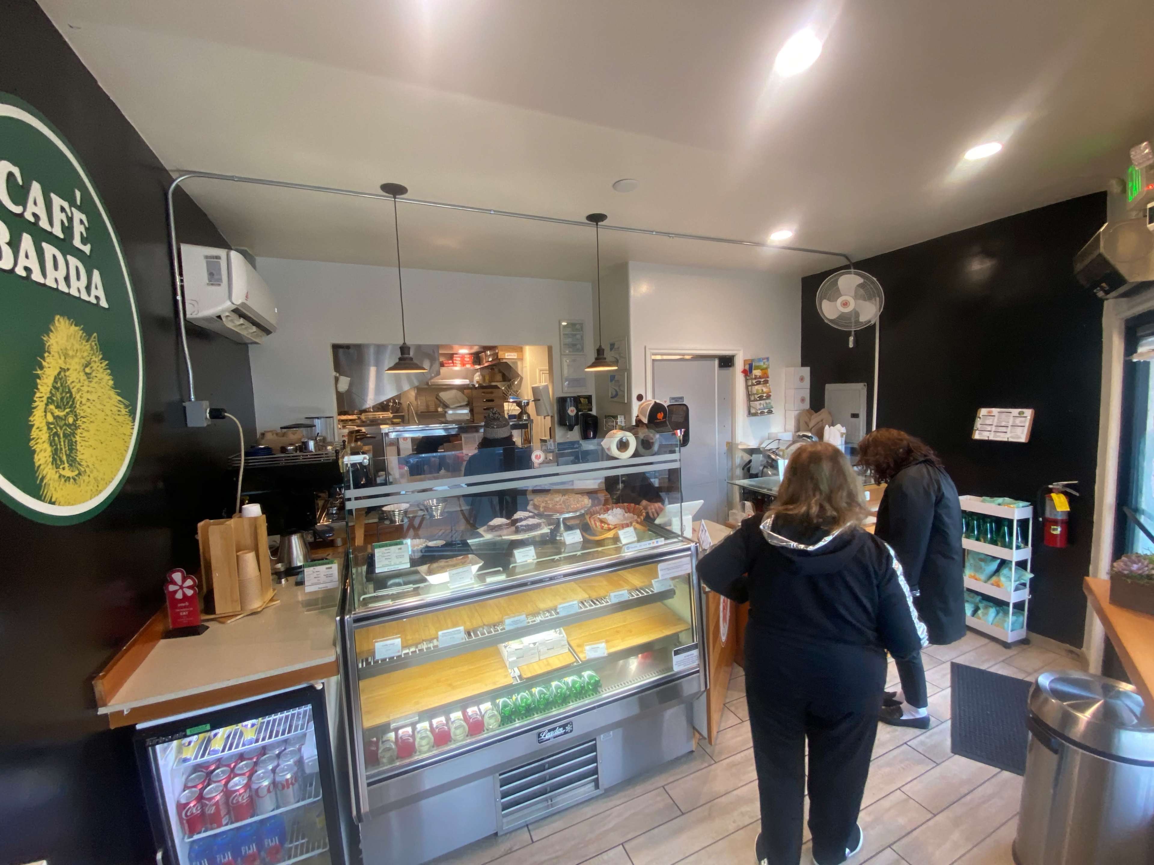 The interior of a cafe with a glass display case filled with food items and two patrons ordering at the counter.