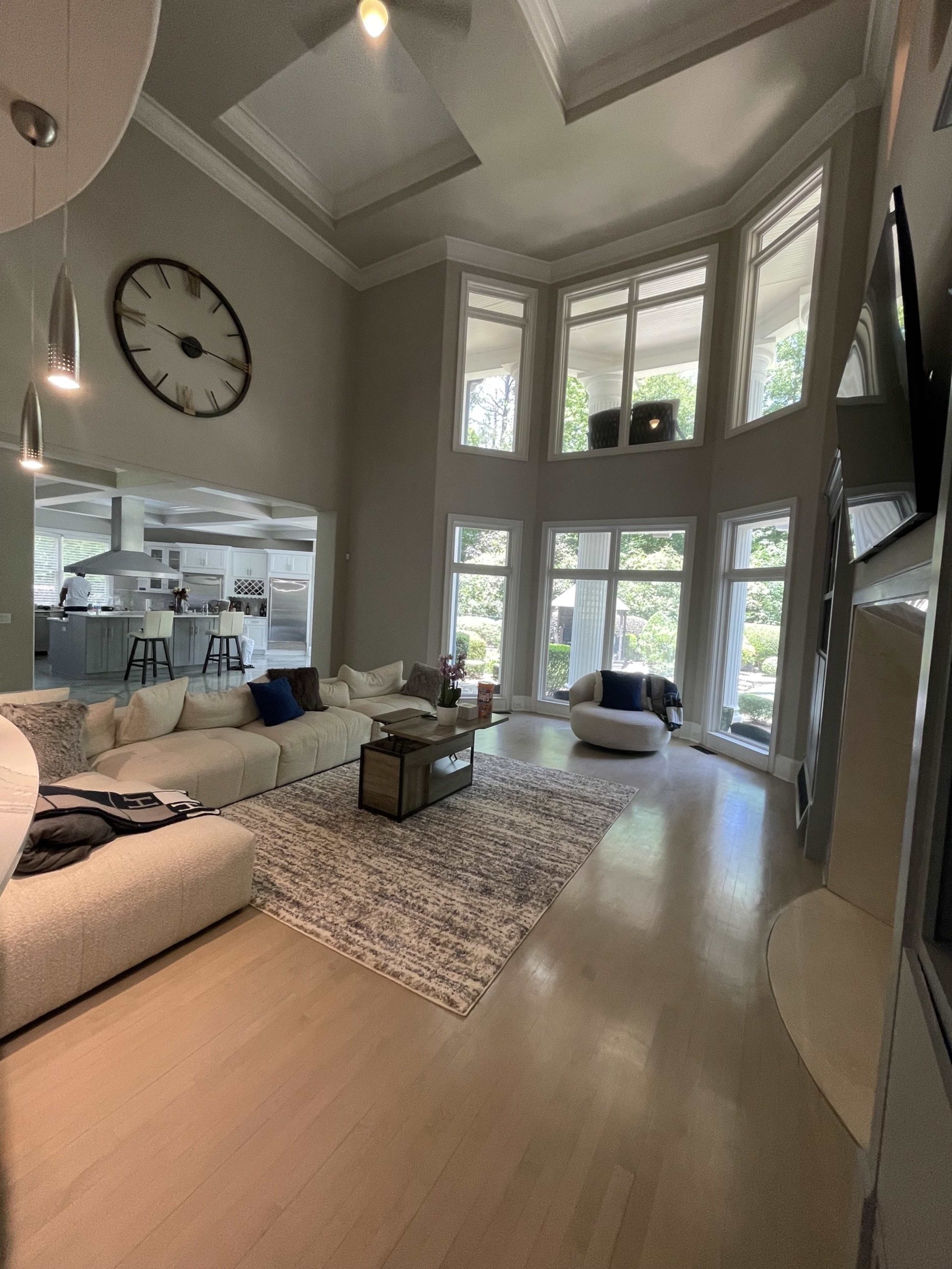 The image shows a spacious living room with large windows, a sectional sofa, a coffee table, and a view of a kitchen in the background.