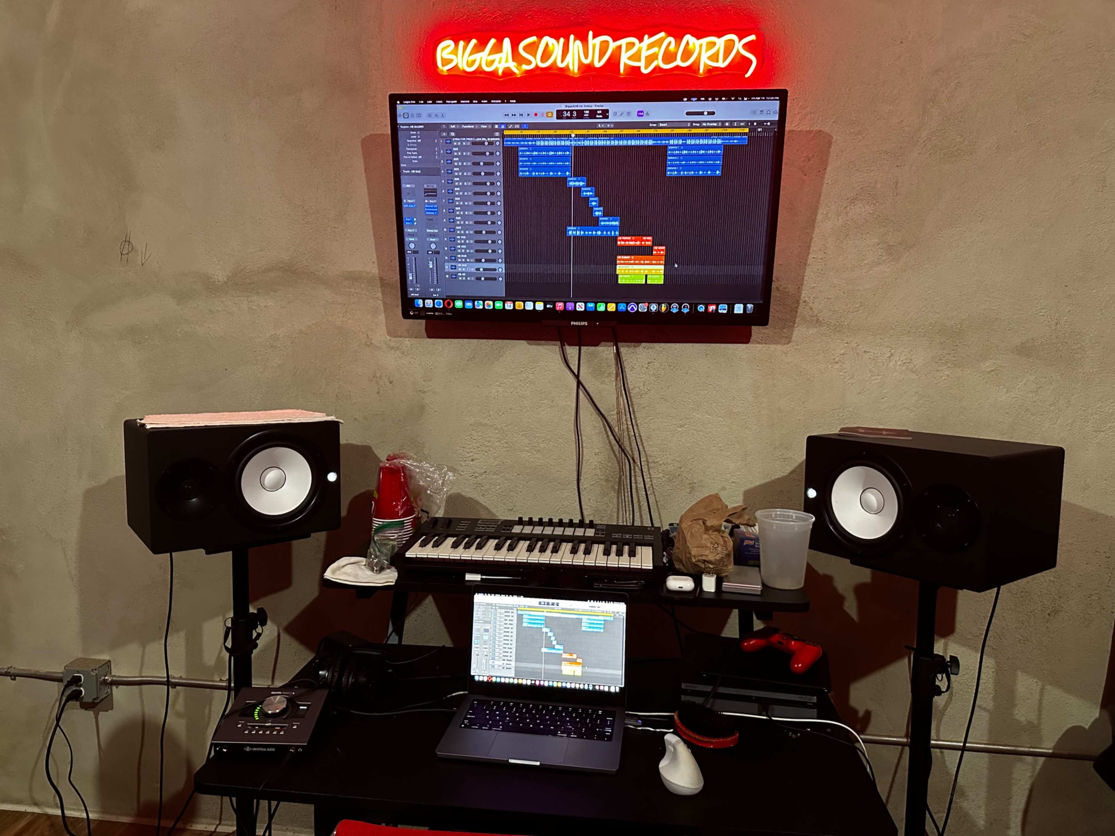 A music production setup with a computer screen displaying a digital audio workstation, two studio monitors, a keyboard, and various equipment on a dark table under a brightly lit sign.