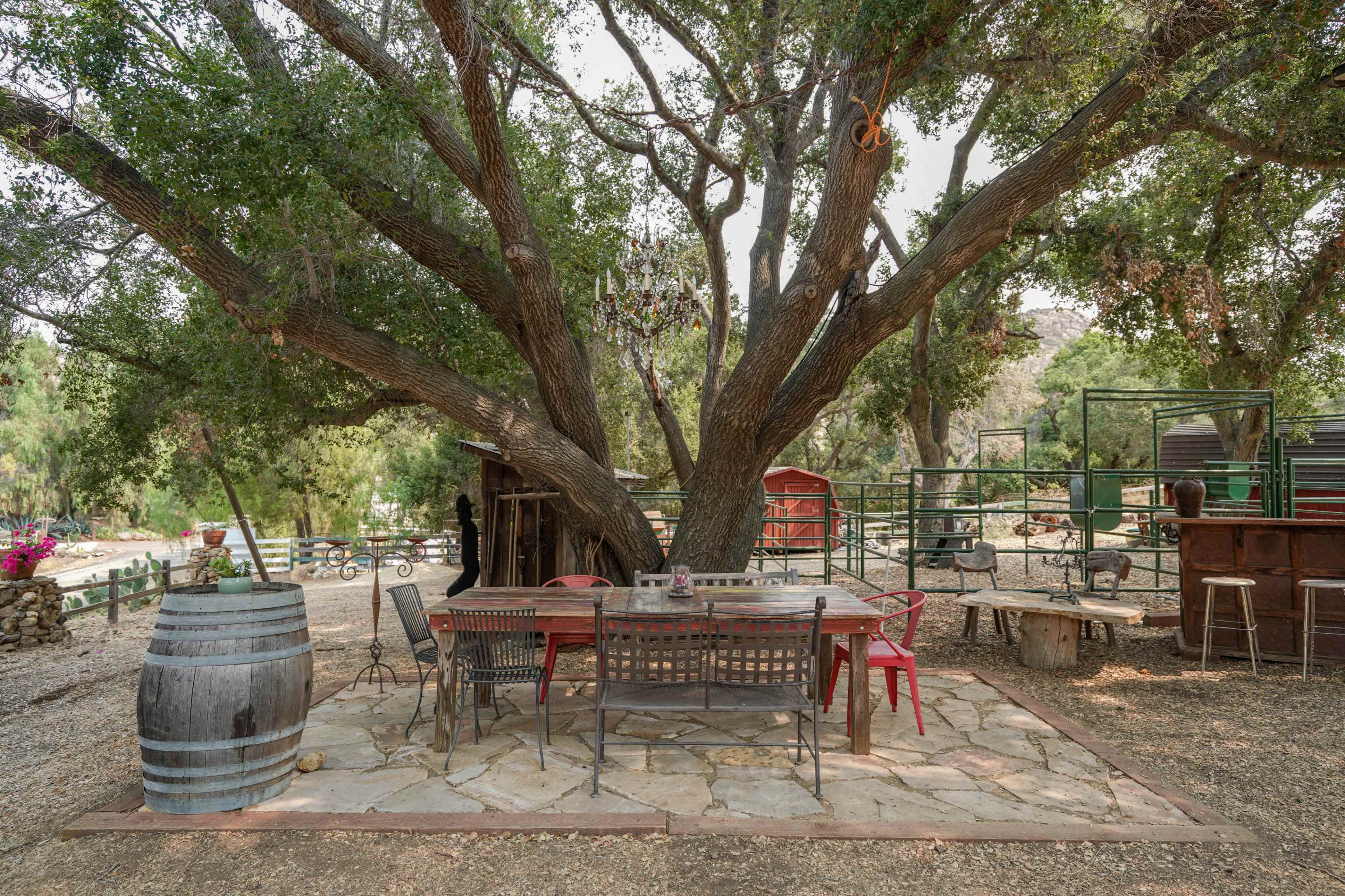 A stone patio with a large table and chairs is shaded by a sprawling tree, surrounded by rustic decor and green fencing.
