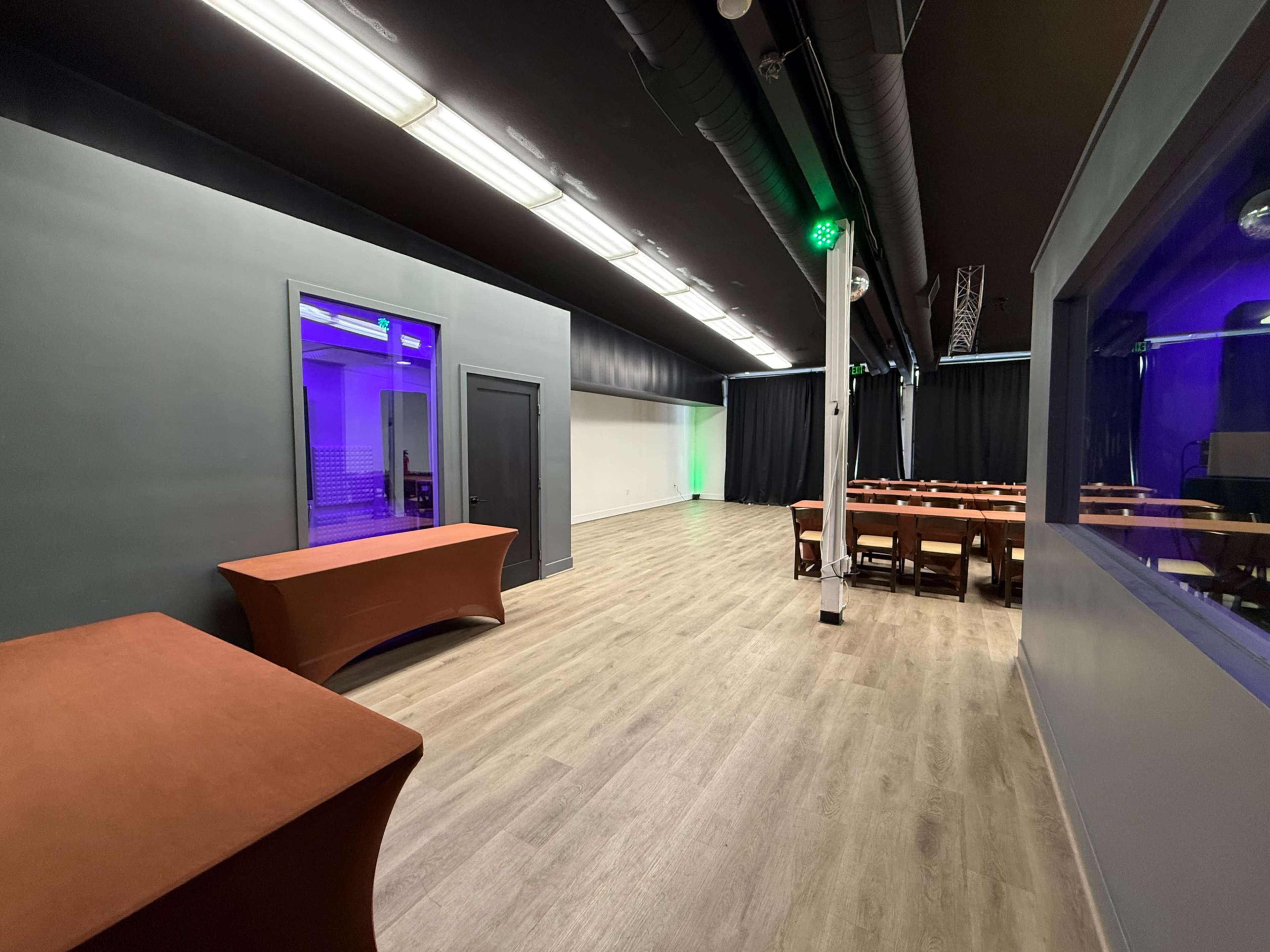 The image shows an empty room with wooden flooring, featuring a stage area with curtains and a table on the left side.