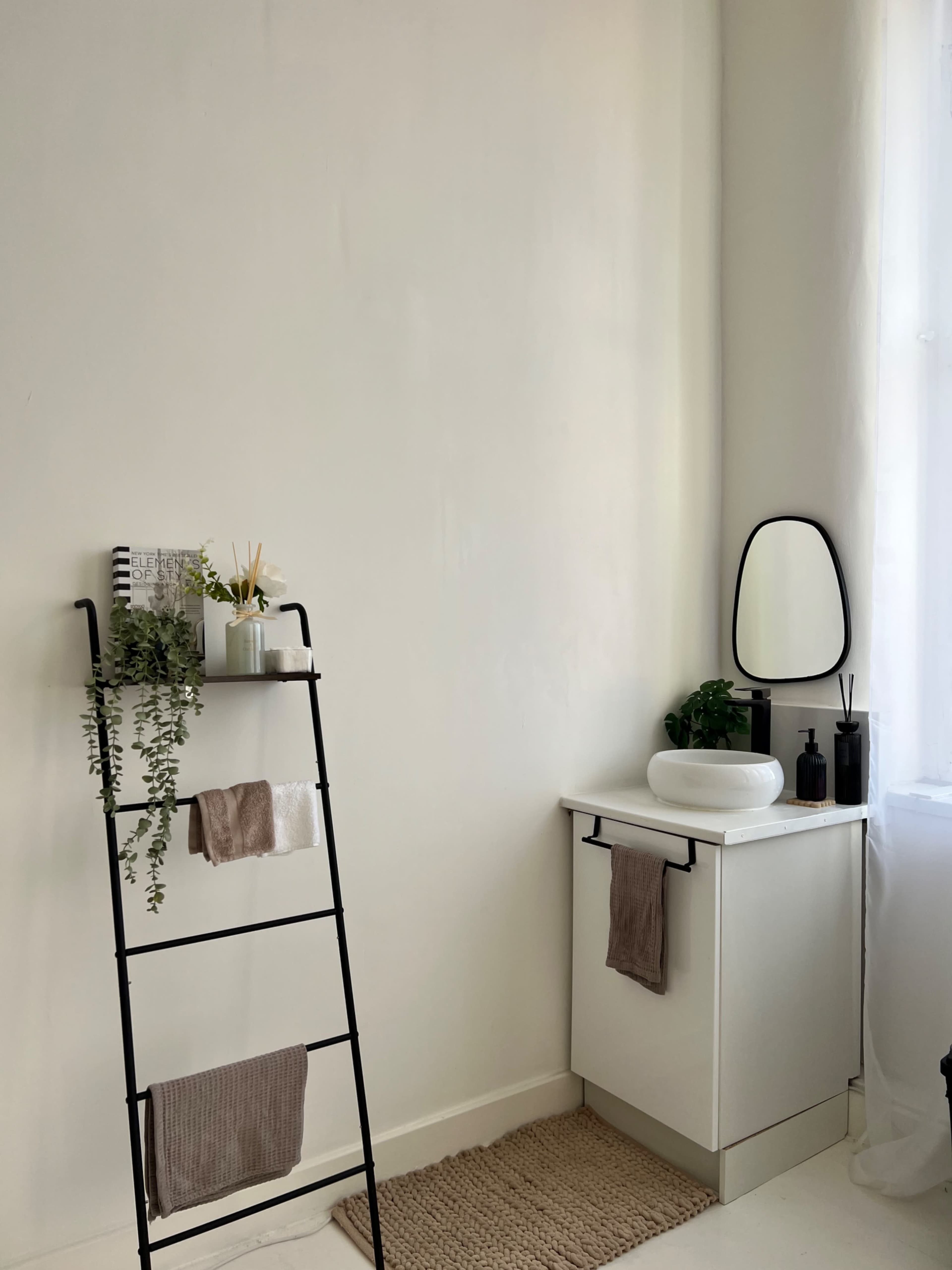The image shows a minimalist bathroom setting featuring a black ladder shelf with plants and towels, a white sink on a cabinet, and a mirror near a window.