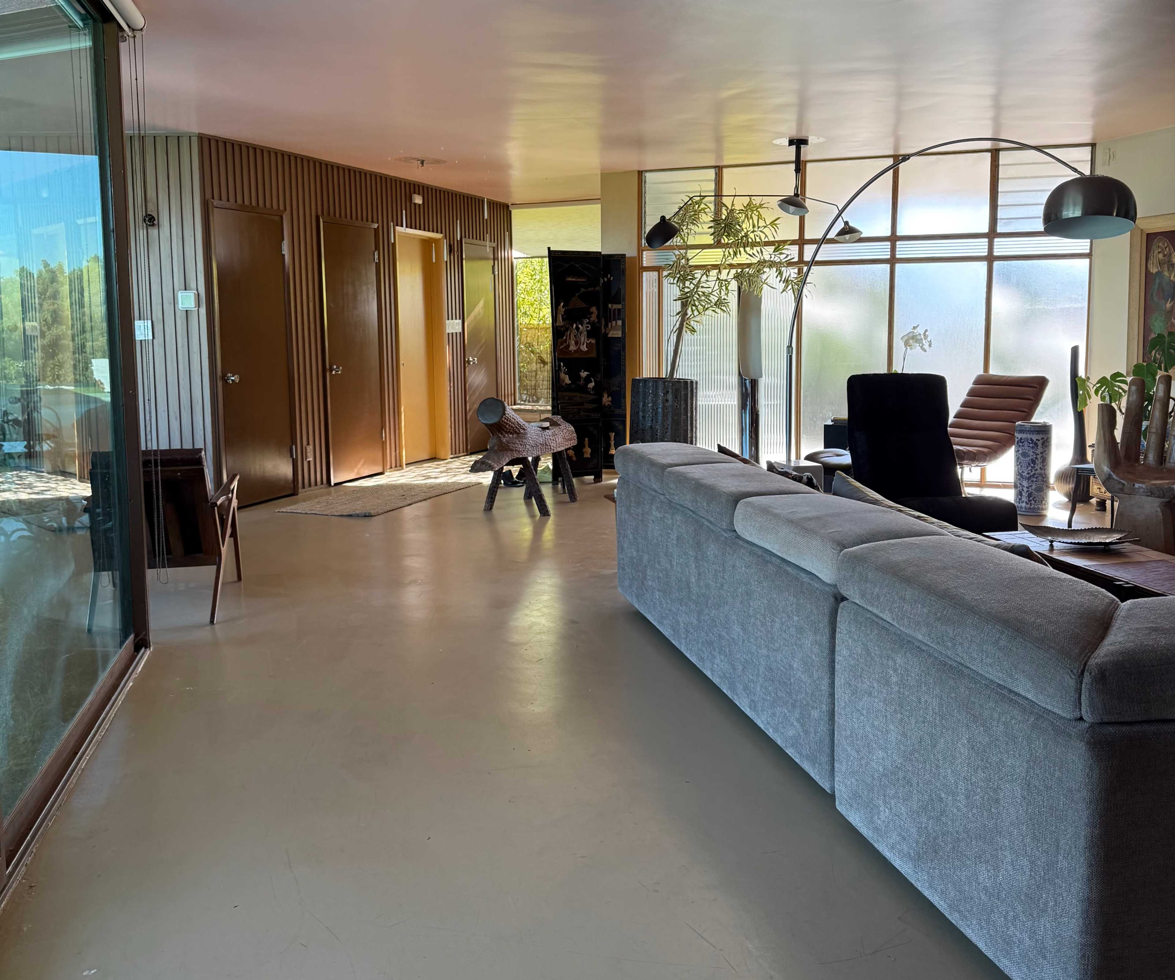 The image shows a modern living space with a gray sofa, large windows, and wooden doors leading to different rooms.