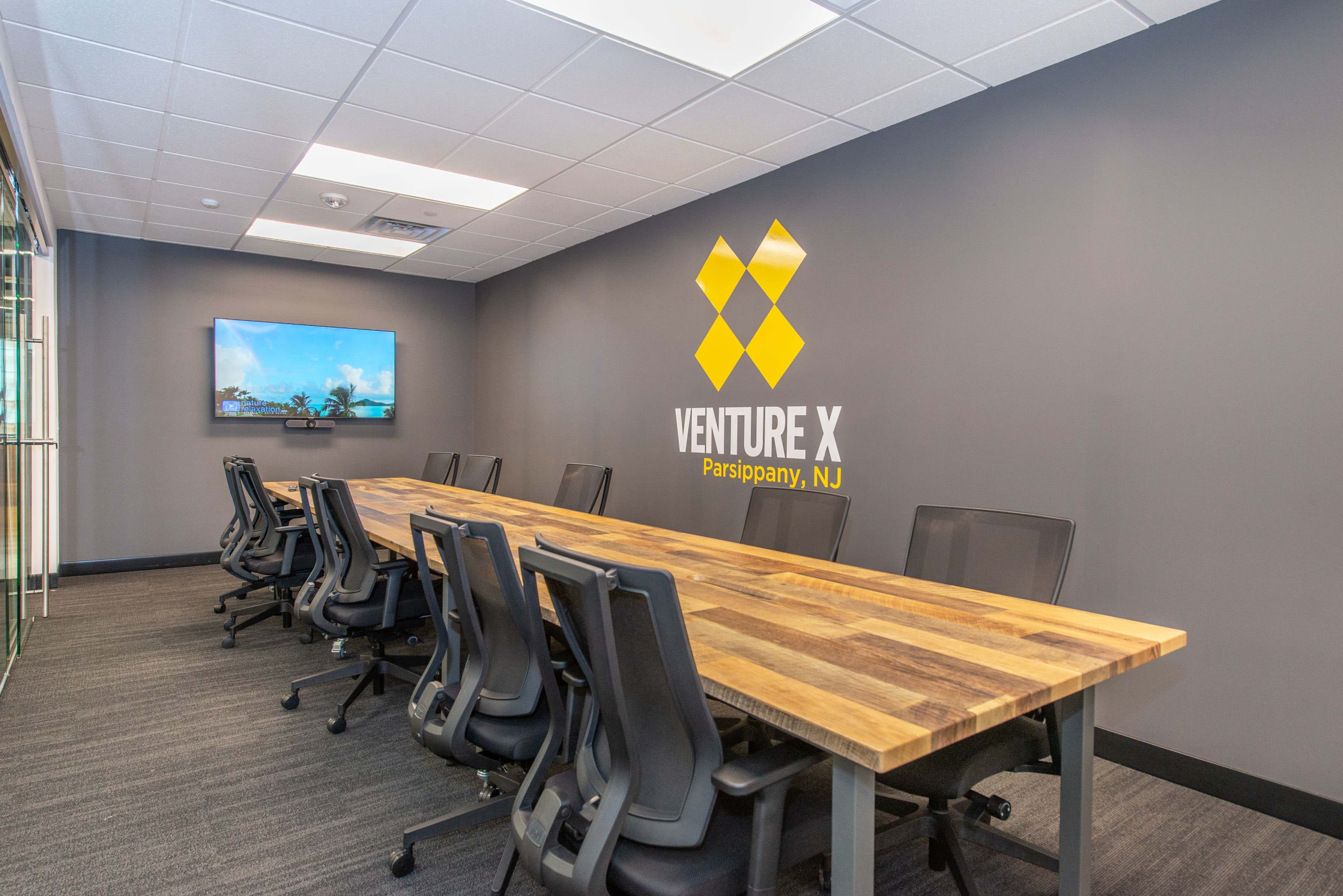 A long wooden conference table with black chairs is set up in a meeting room featuring a TV and a wall with a yellow logo for Venture X in Parsippany, NJ.