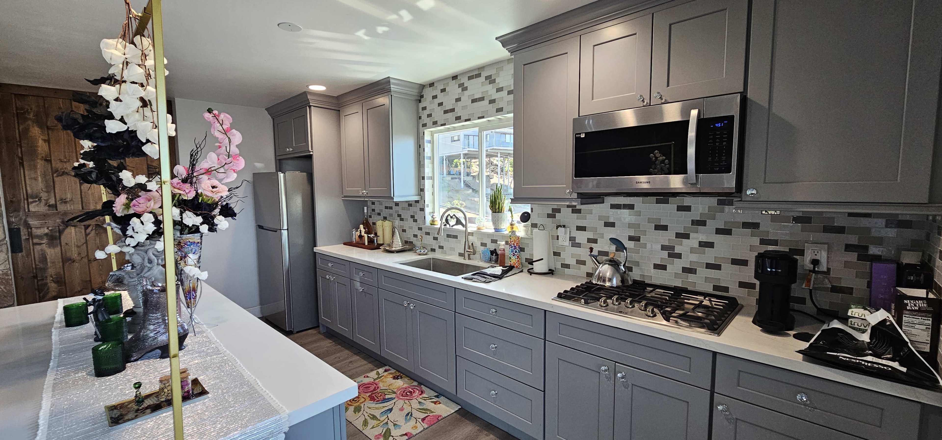 The image shows a modern kitchen featuring gray cabinetry, a stainless steel microwave, a stove, and decorative flowers on the countertop.