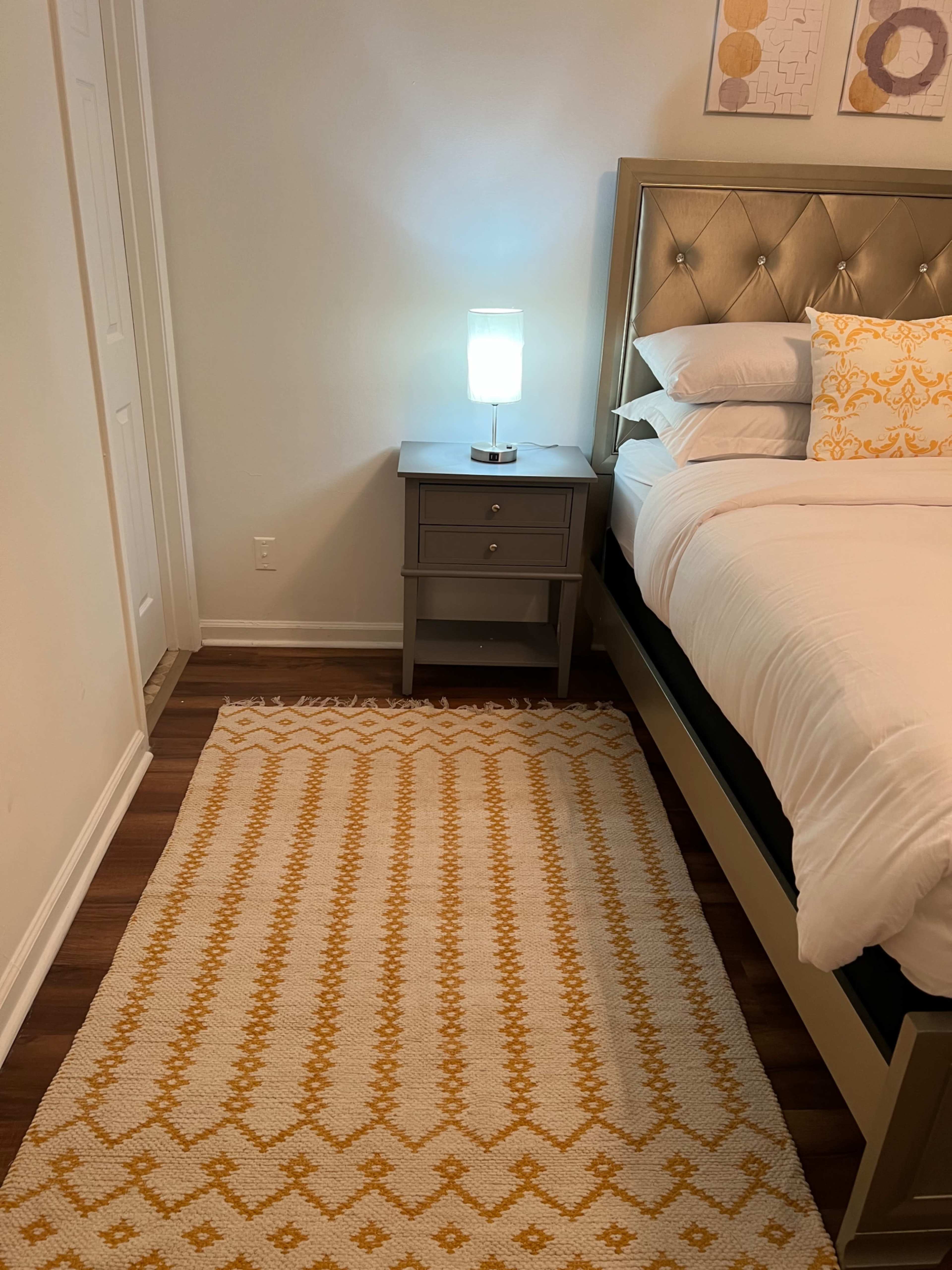 A neatly made bed with white linens, a gray nightstand with a lamp, and a patterned yellow and white rug on a wooden floor.