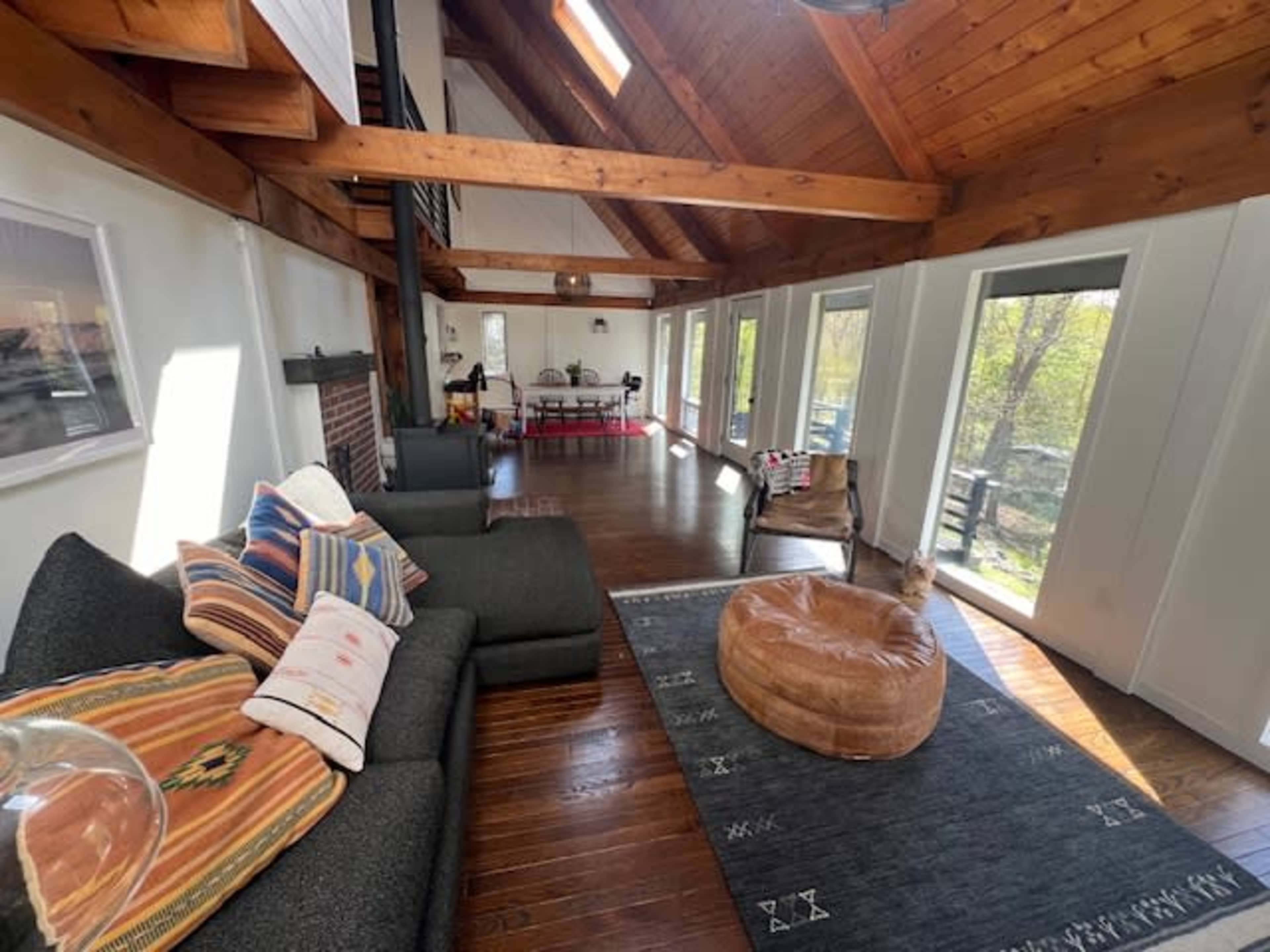 A spacious living area with a dark sofa, a round tan pouf, and large windows that provide views of an outdoor space.
