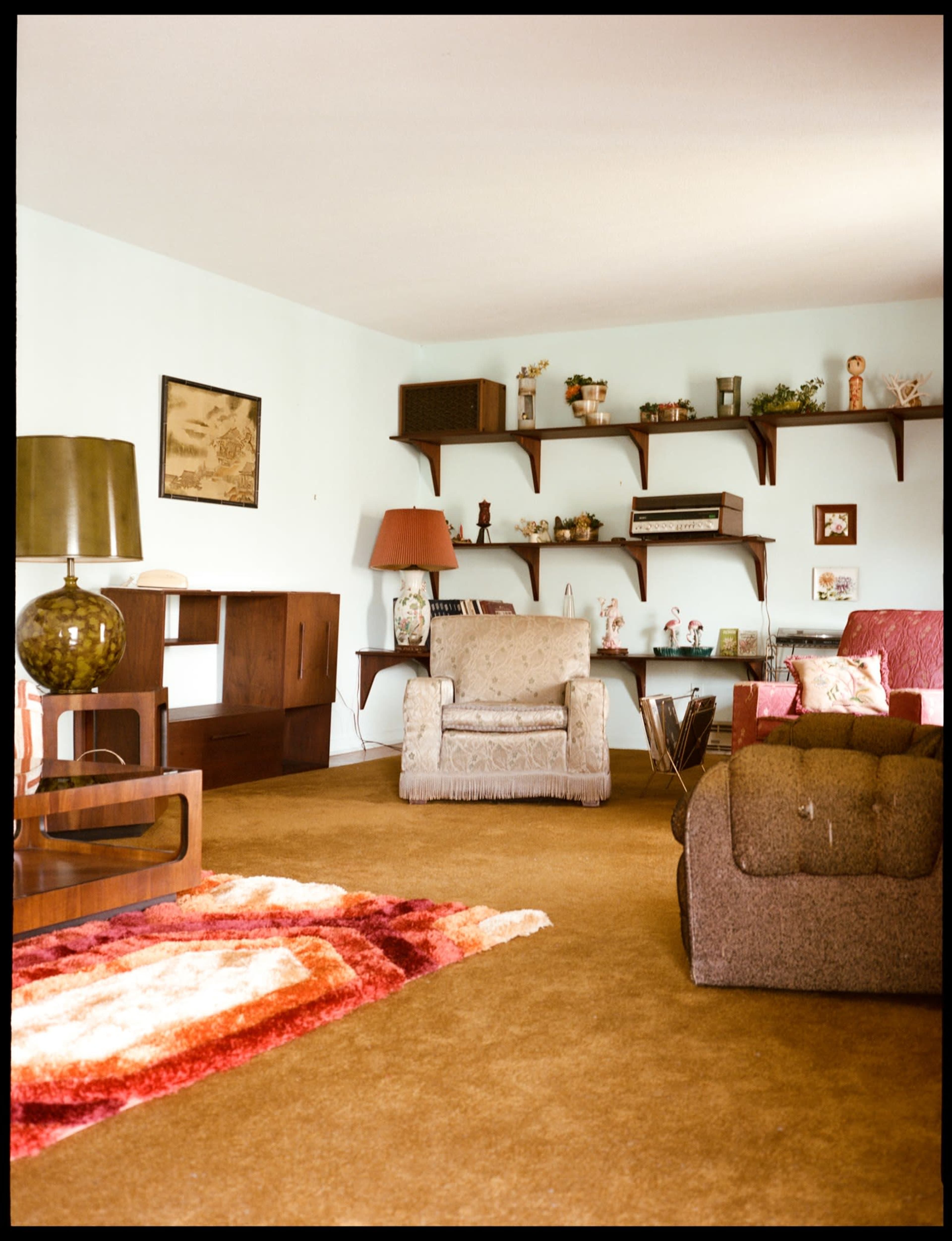 The image shows a retro living room with two upholstered chairs, a wooden shelf displaying various items, and a patterned rug on a carpeted floor.