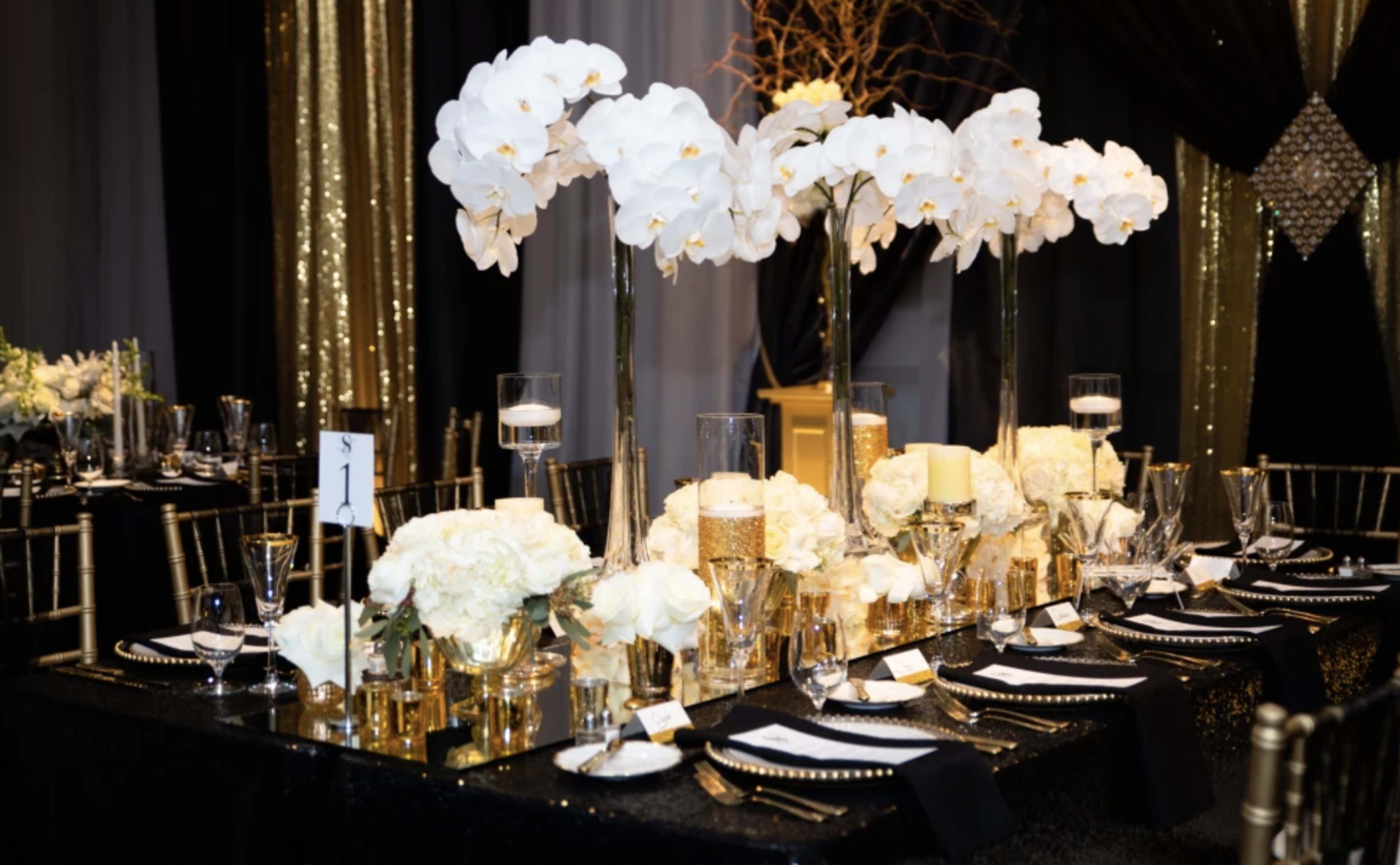 A elegantly set dining table features tall white orchid centerpieces, gold accents, and crystal glassware against a backdrop of black and gold decor.