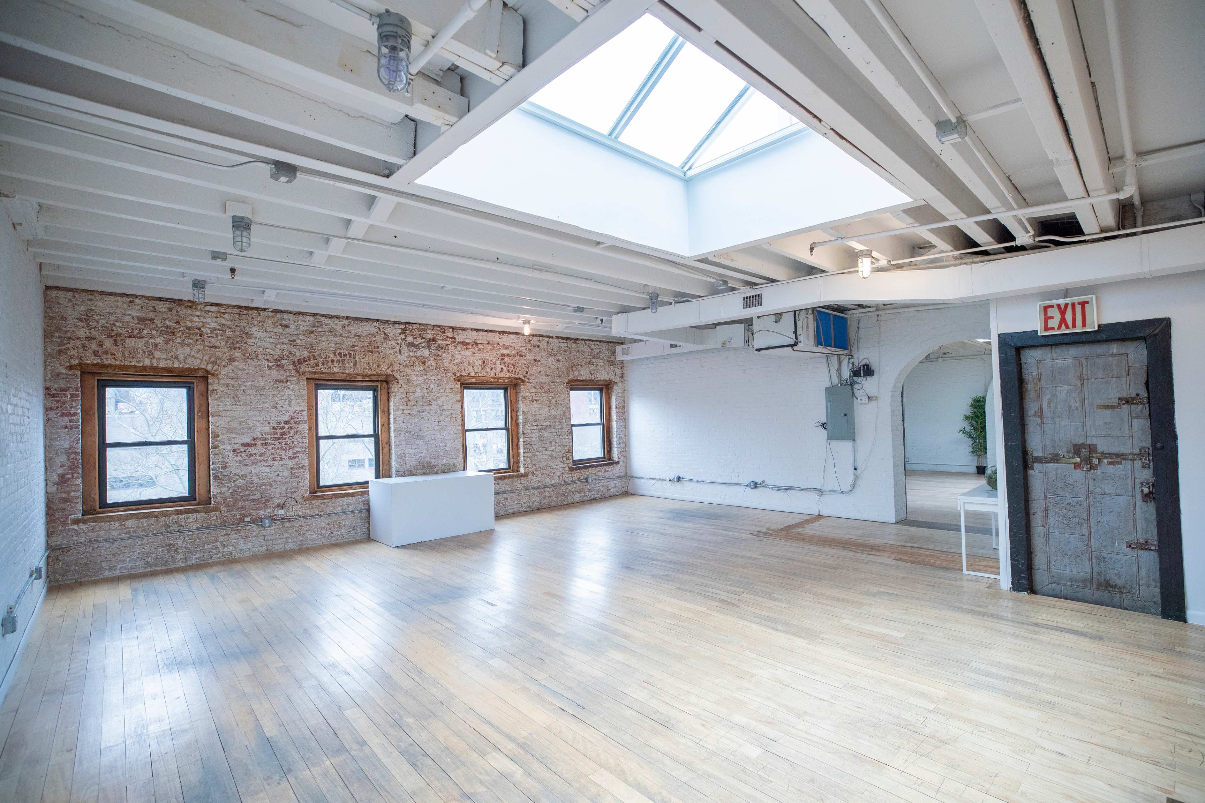 The image shows a spacious, well-lit interior with exposed brick walls, wooden floors, and large windows, featuring a skylight overhead.