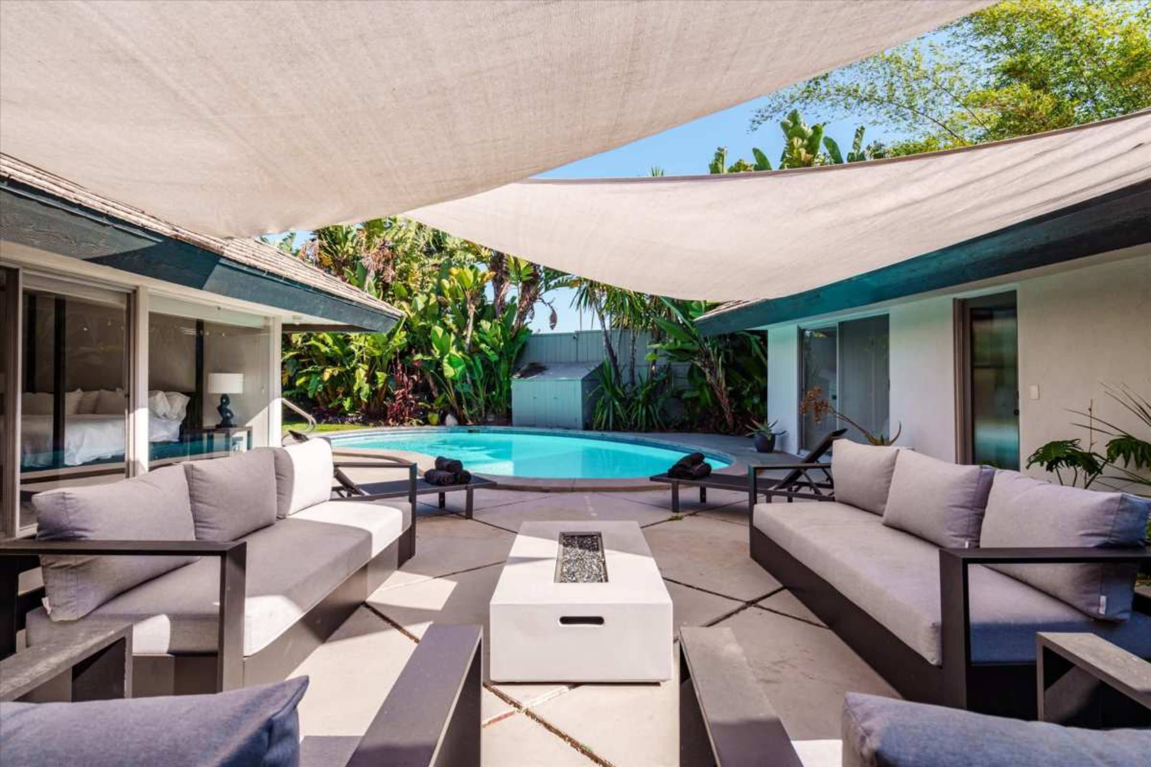 A covered outdoor seating area surrounds a swimming pool, with lush greenery in the background.