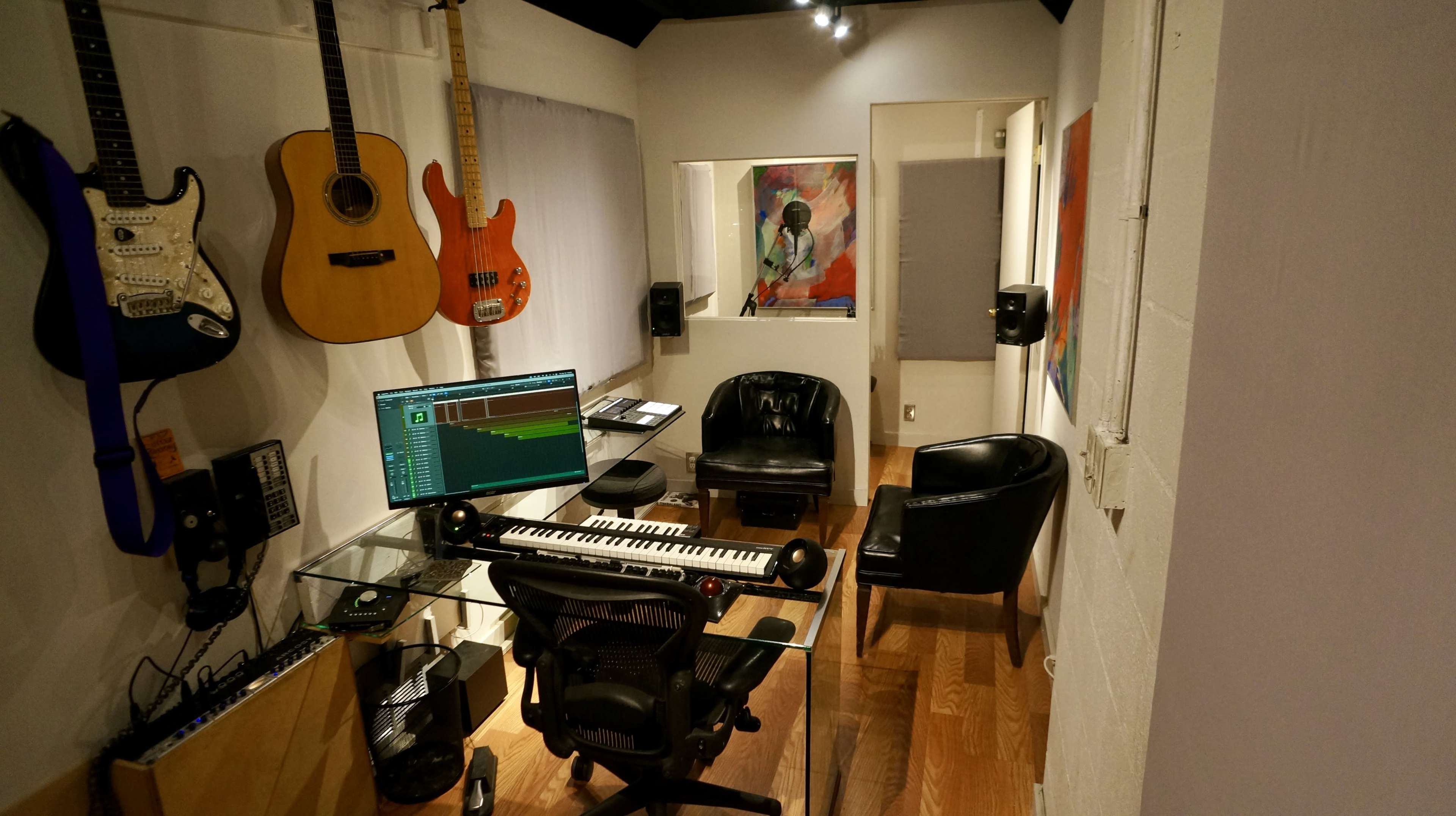 A small music studio features a glass-enclosed vocal booth, an array of guitars mounted on the wall, and a desk with a computer and keyboard.