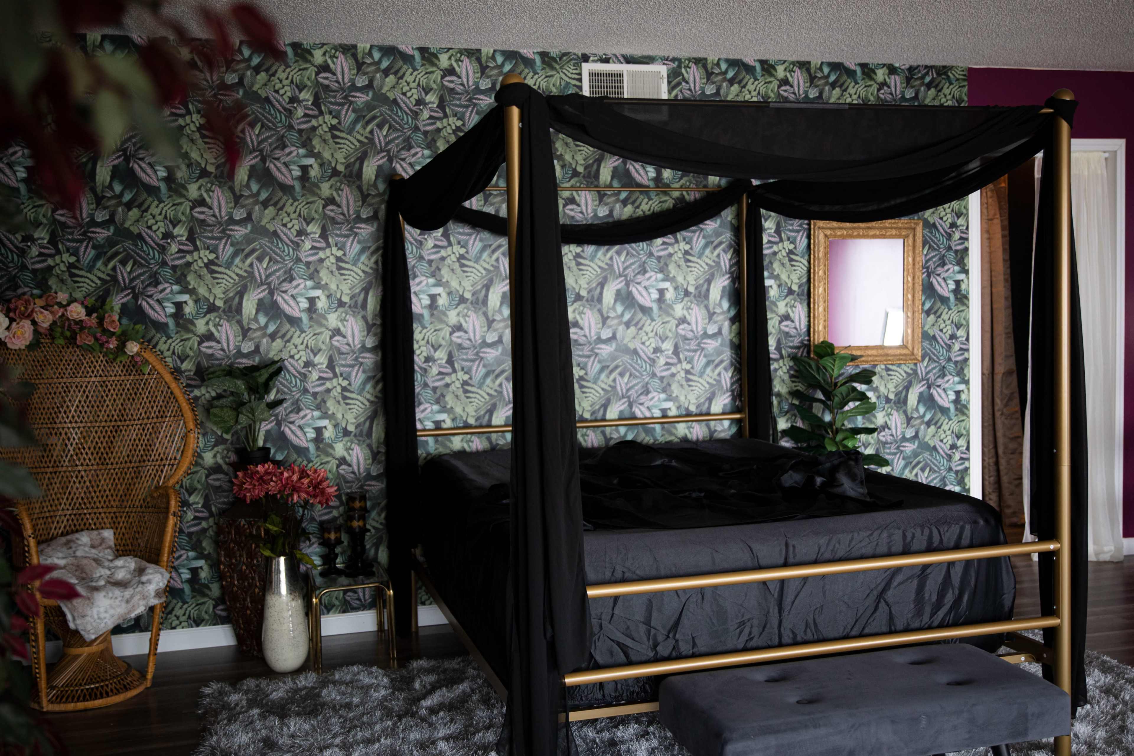 A bedroom with a canopy bed draped in black fabric, surrounded by leafy wallpaper and a decorative wicker chair.